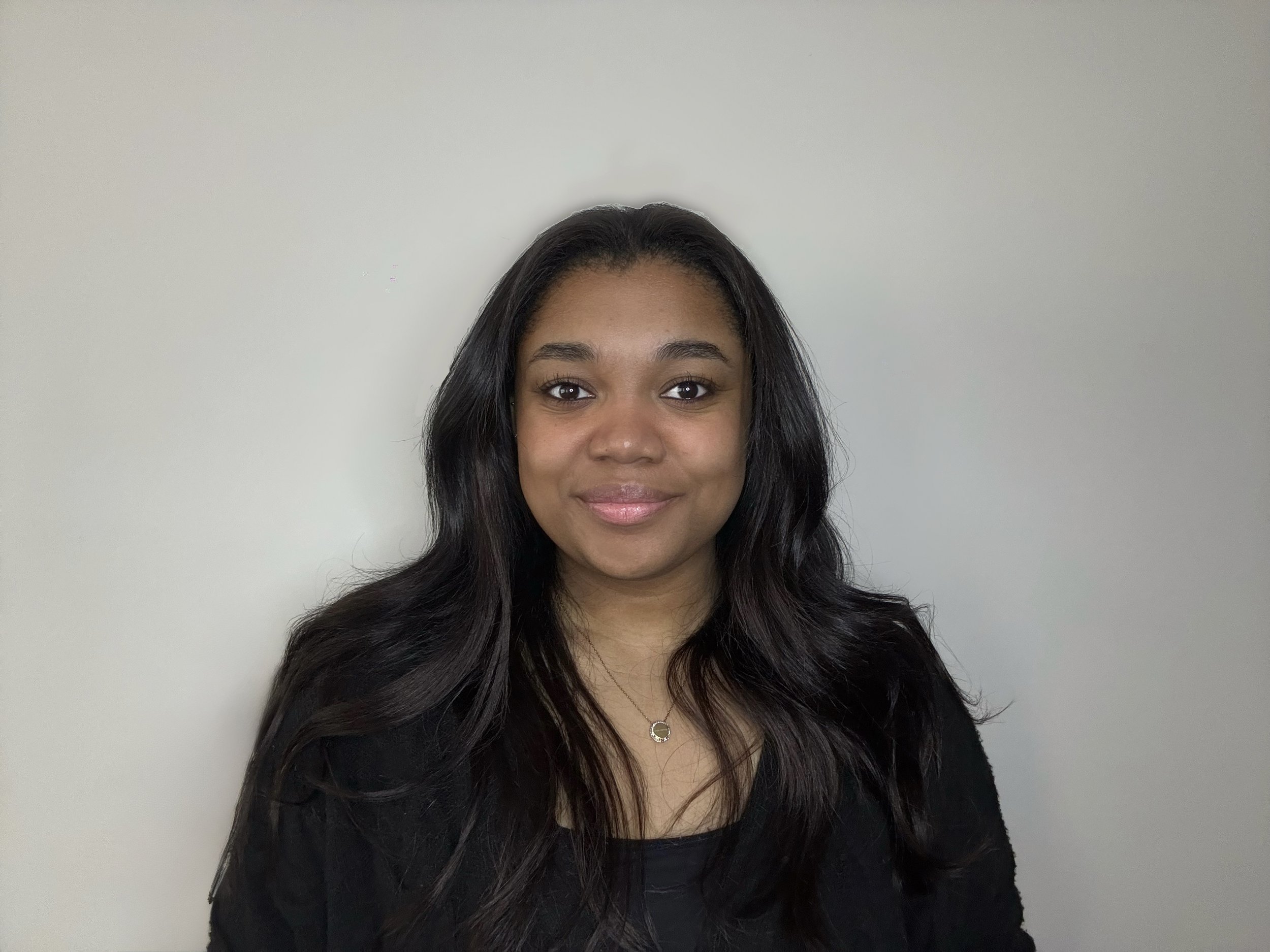 A young woman with long, wavy black hair, smiling, wearing a black top and a necklace with a small pendant, standing against a plain light-colored wall.