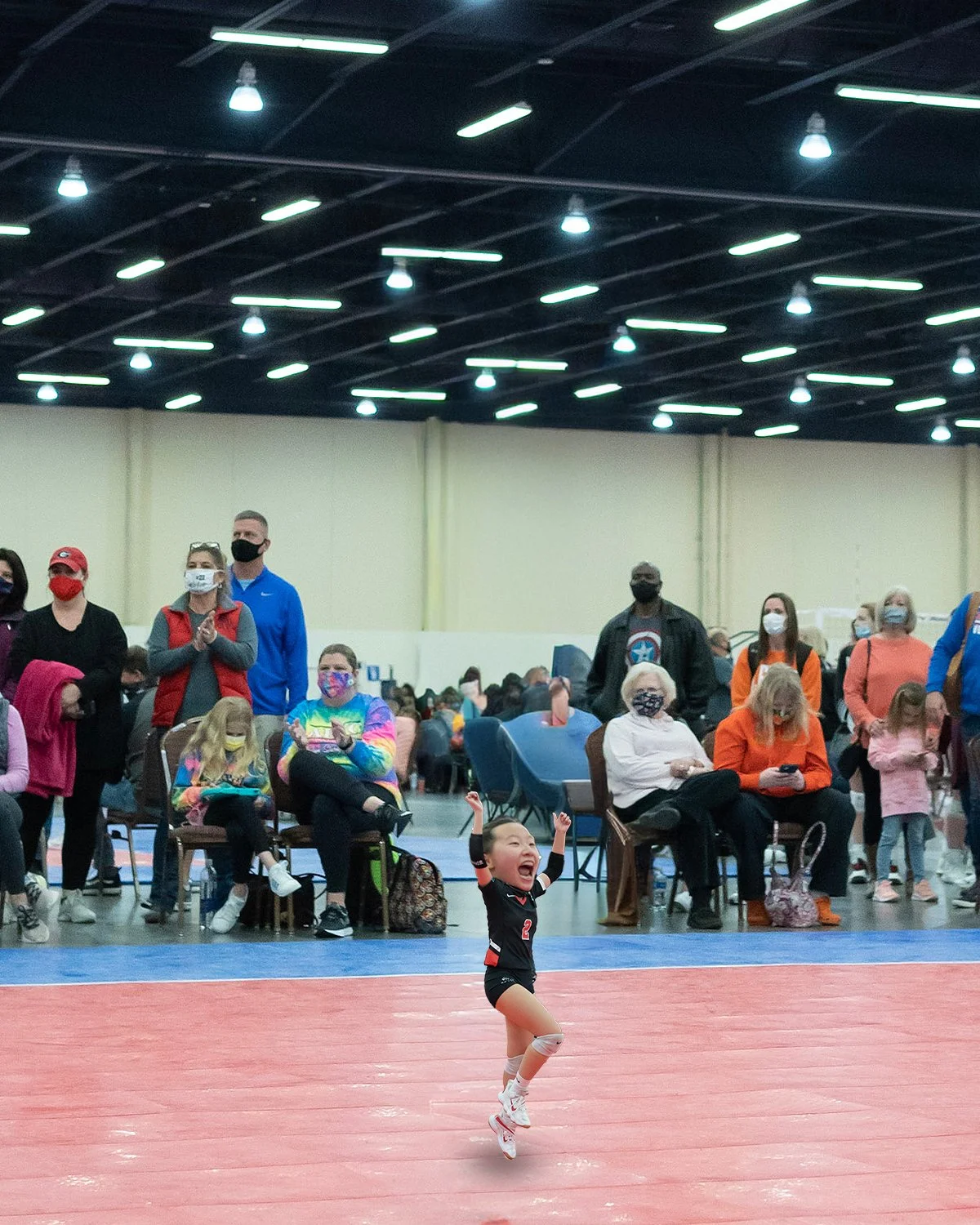 A young girl celebrating on an indoor volleyball court with a crowd of spectators watching in the background.