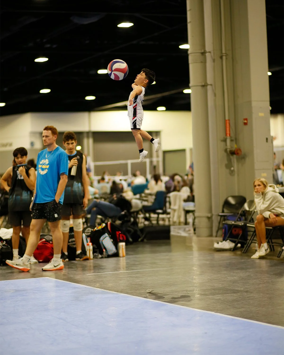 A young boy in a white sports outfit jumps in the air and hits a volleyball at an indoor sports arena, with spectators and other players in the background.