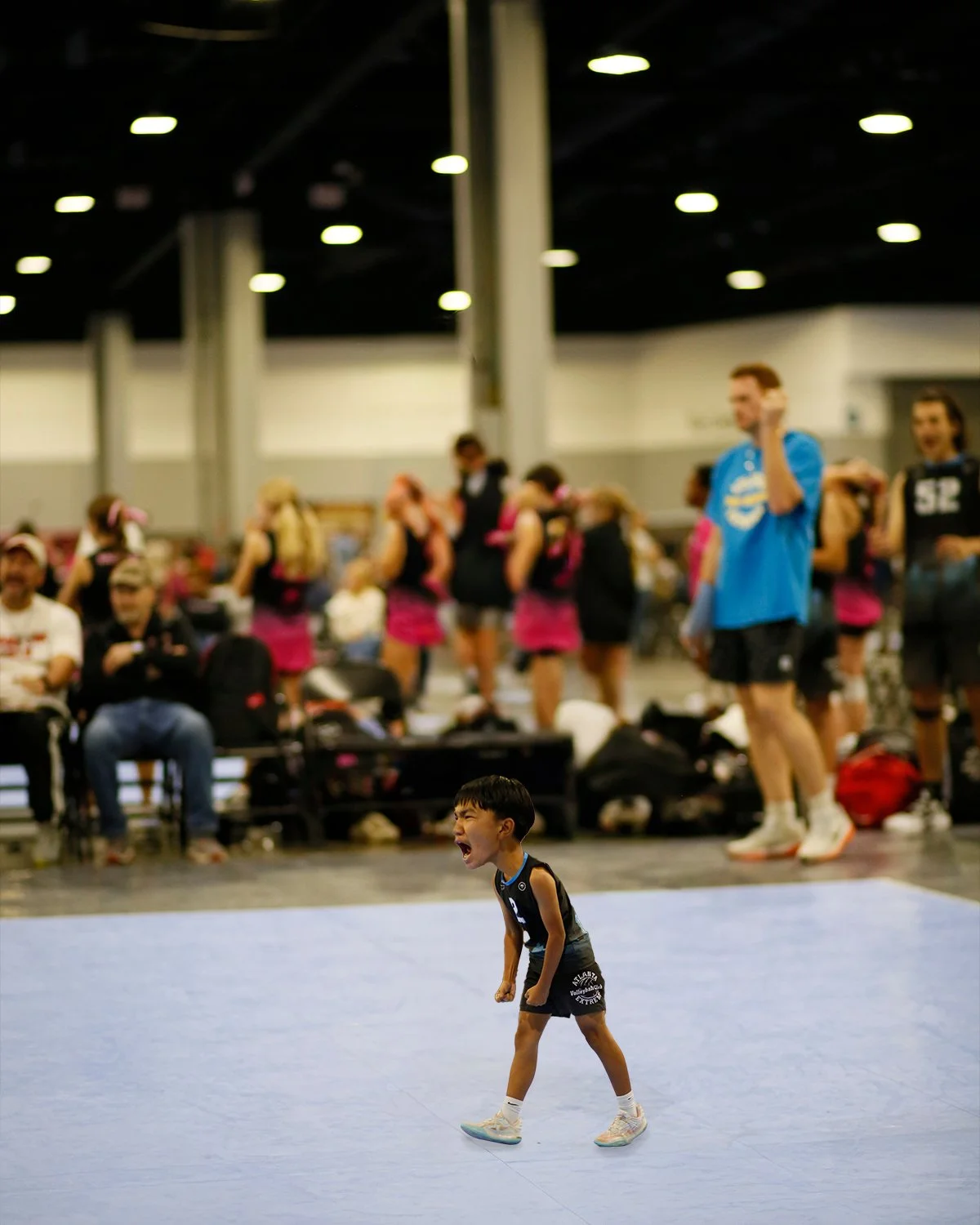 A young boy wearing a black wrestling uniform on a blue wrestling mat, shouting or yelling with emotion. In the background, there are people including women in pink and black uniforms, and some seated and standing, with a large indoor space and overh