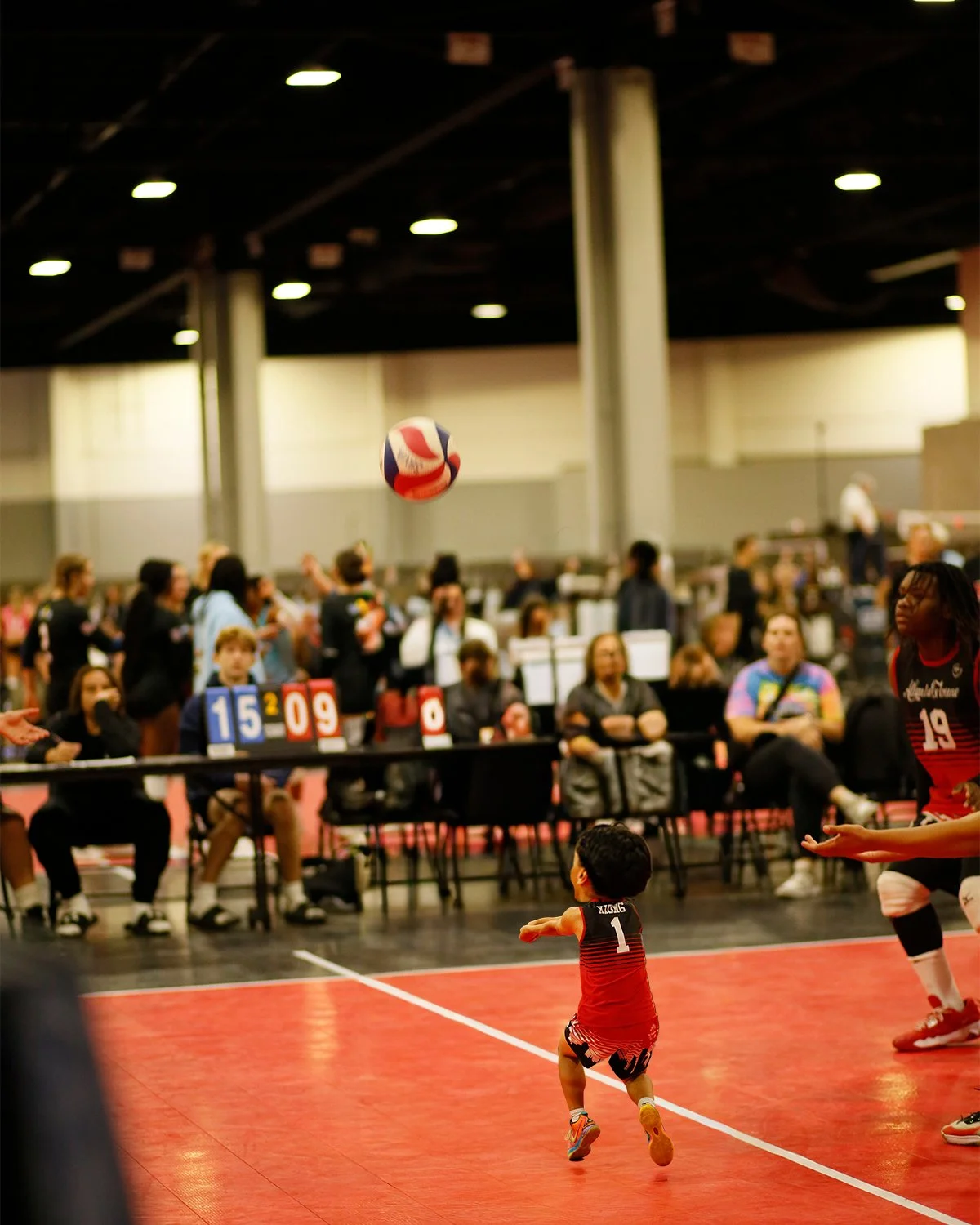 A young child playing volleyball in an indoor court during a game, with spectators and players in the background.