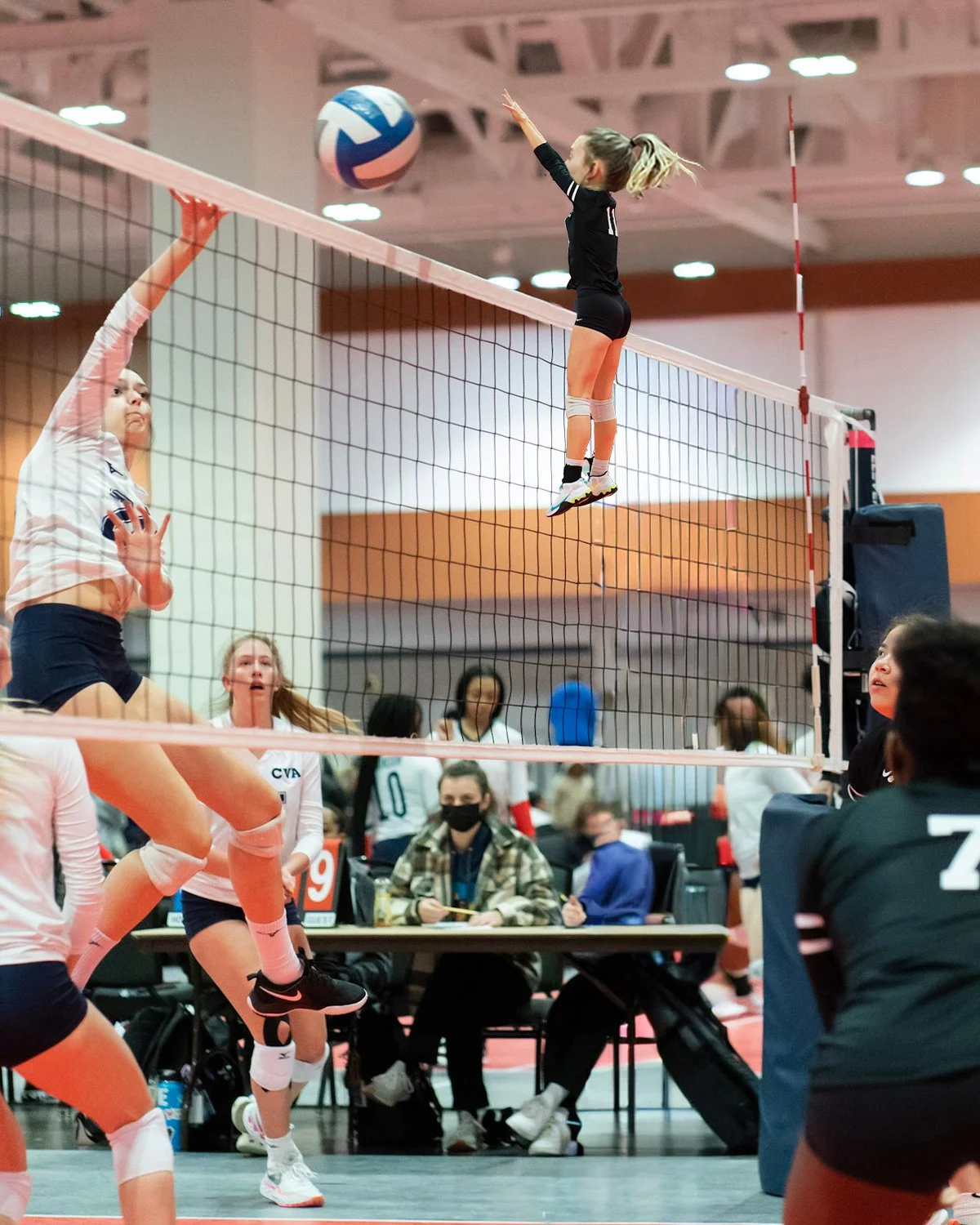 A volleyball game in progress with a female player jumping at the net to spike the ball while an opponent prepares to block. Several players and spectators are visible in the background.