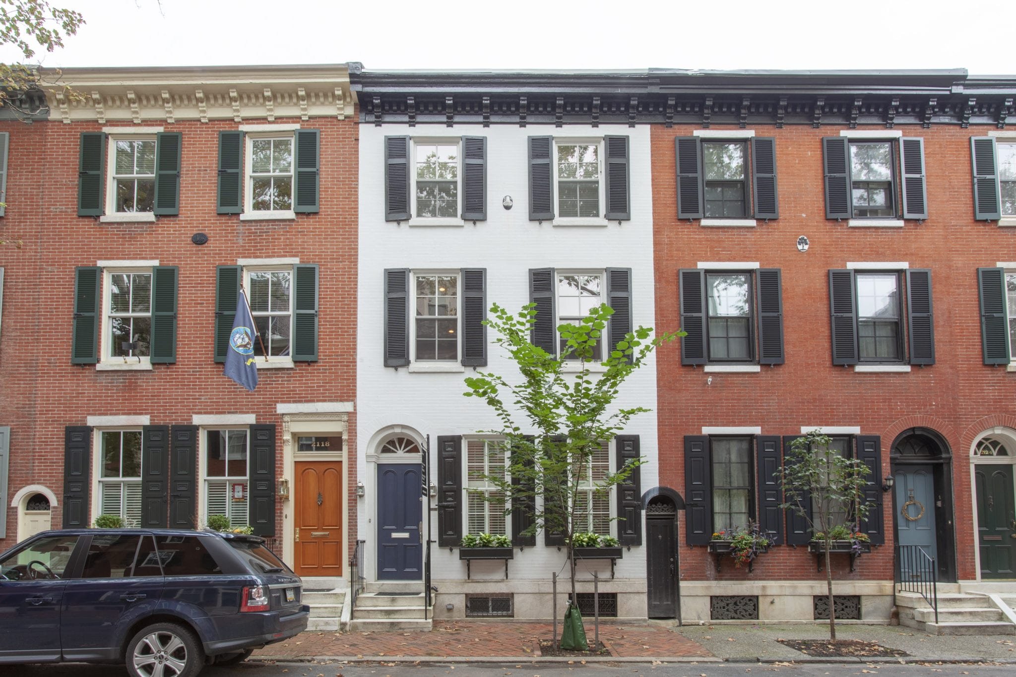 Row of three-story townhouses with brick and white facades, featuring black shuttered windows. A parked SUV is in front, and a small tree is on the sidewalk.