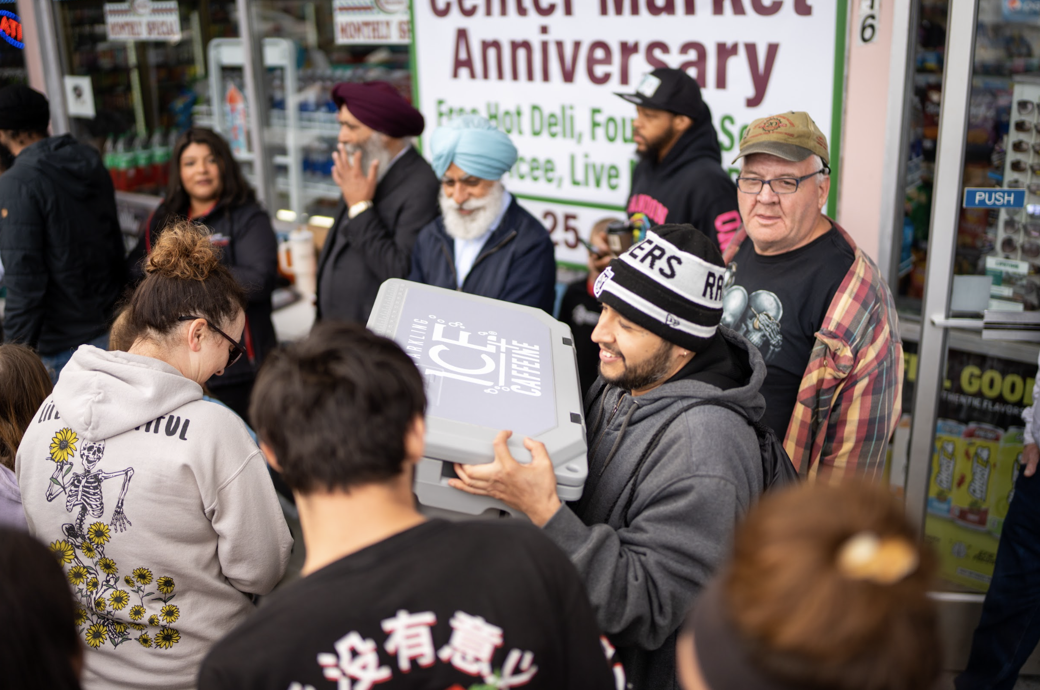 A group of people gathered outside a store celebrating an anniversary, with one person holding a large pizza box and others waiting in line.