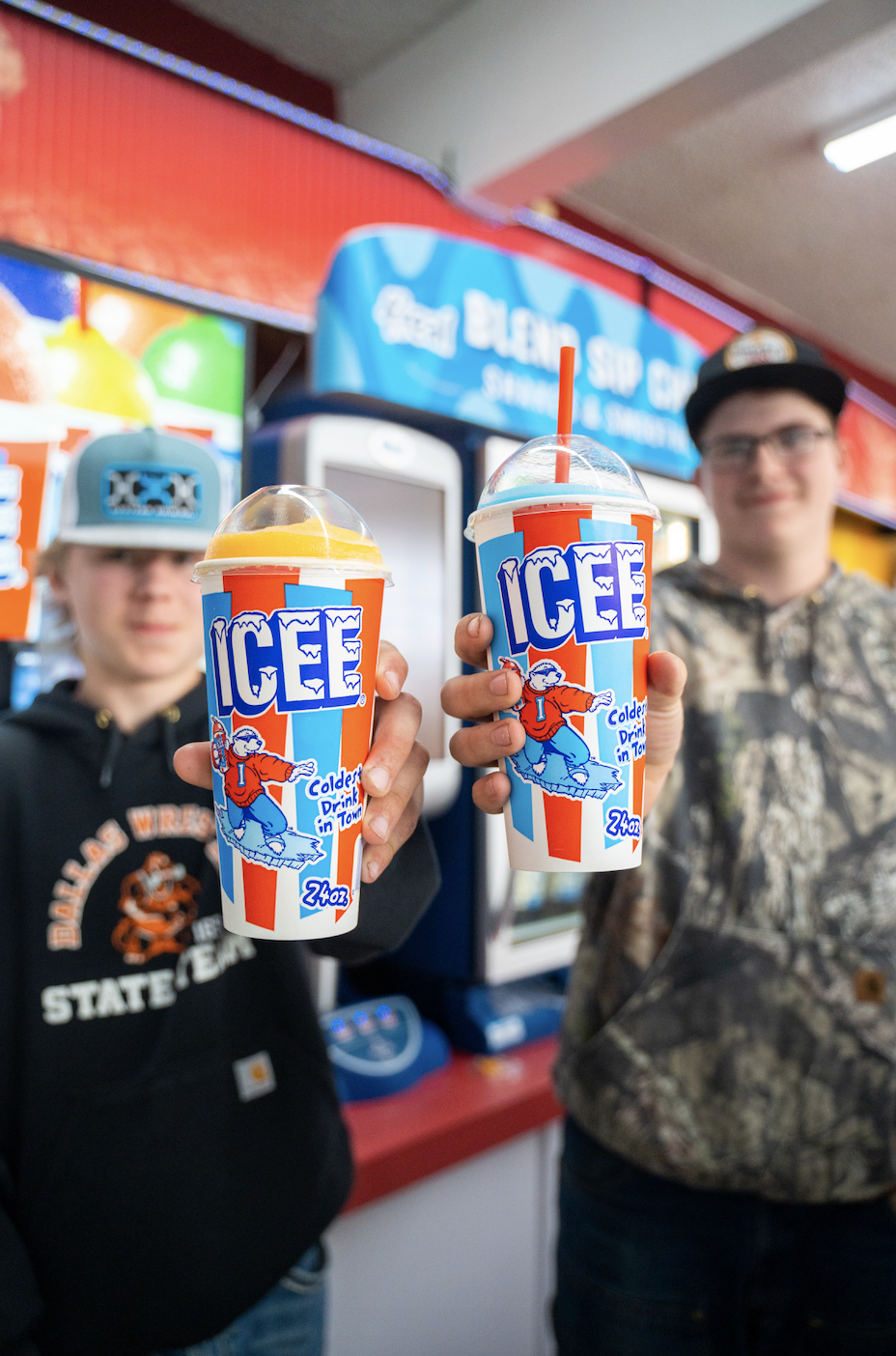Two young men standing at a counter inside an ice cream shop, holding up large cups of icee drinks with colorful striped logos, one with a lemon flavor and the other with a cherry flavor. The background shows bright red, blue, and yellow colors, with