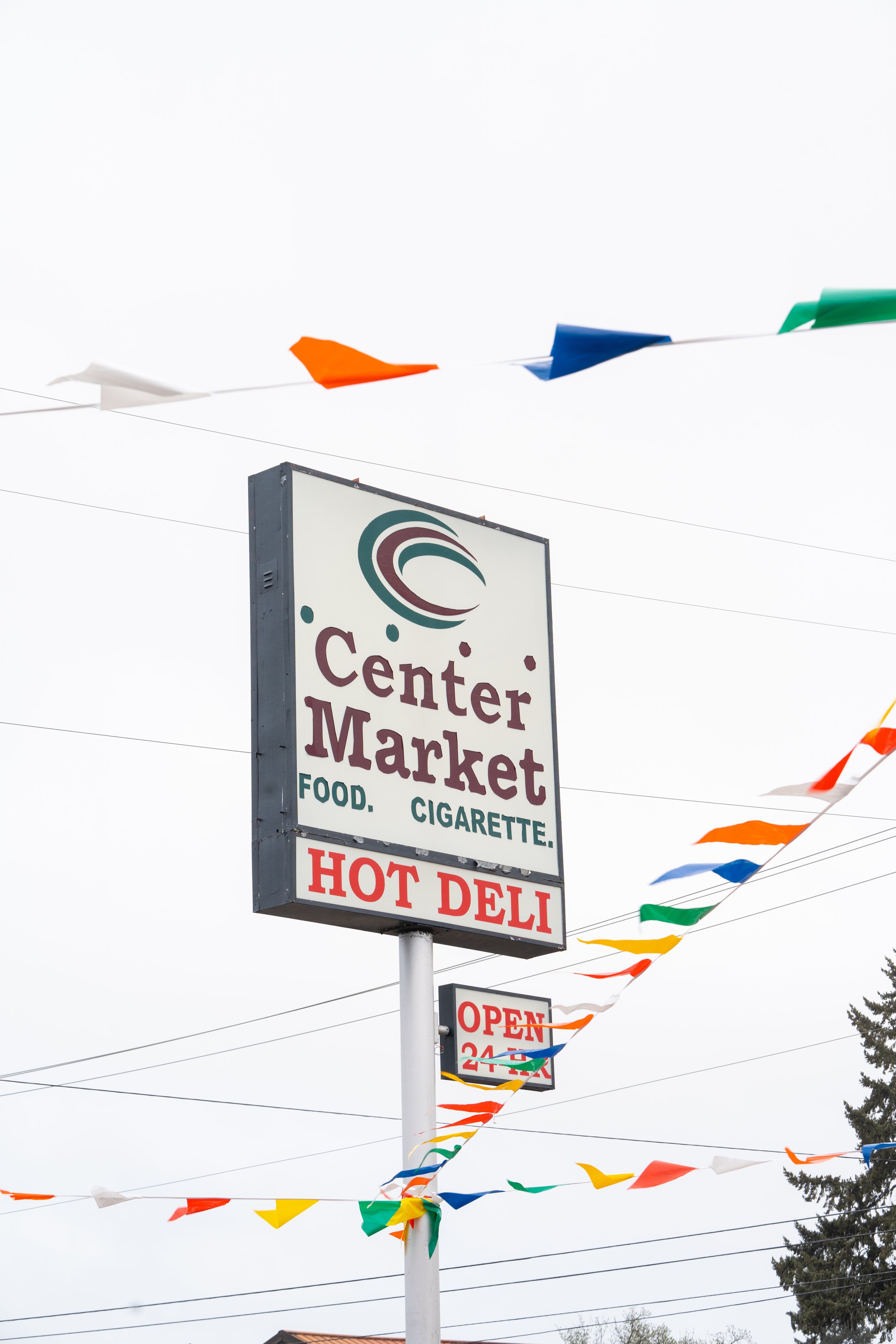 Sign for Center Market, a grocery store that sells food and cigarettes, with colorful flags hanging above and a cloudy sky in the background.