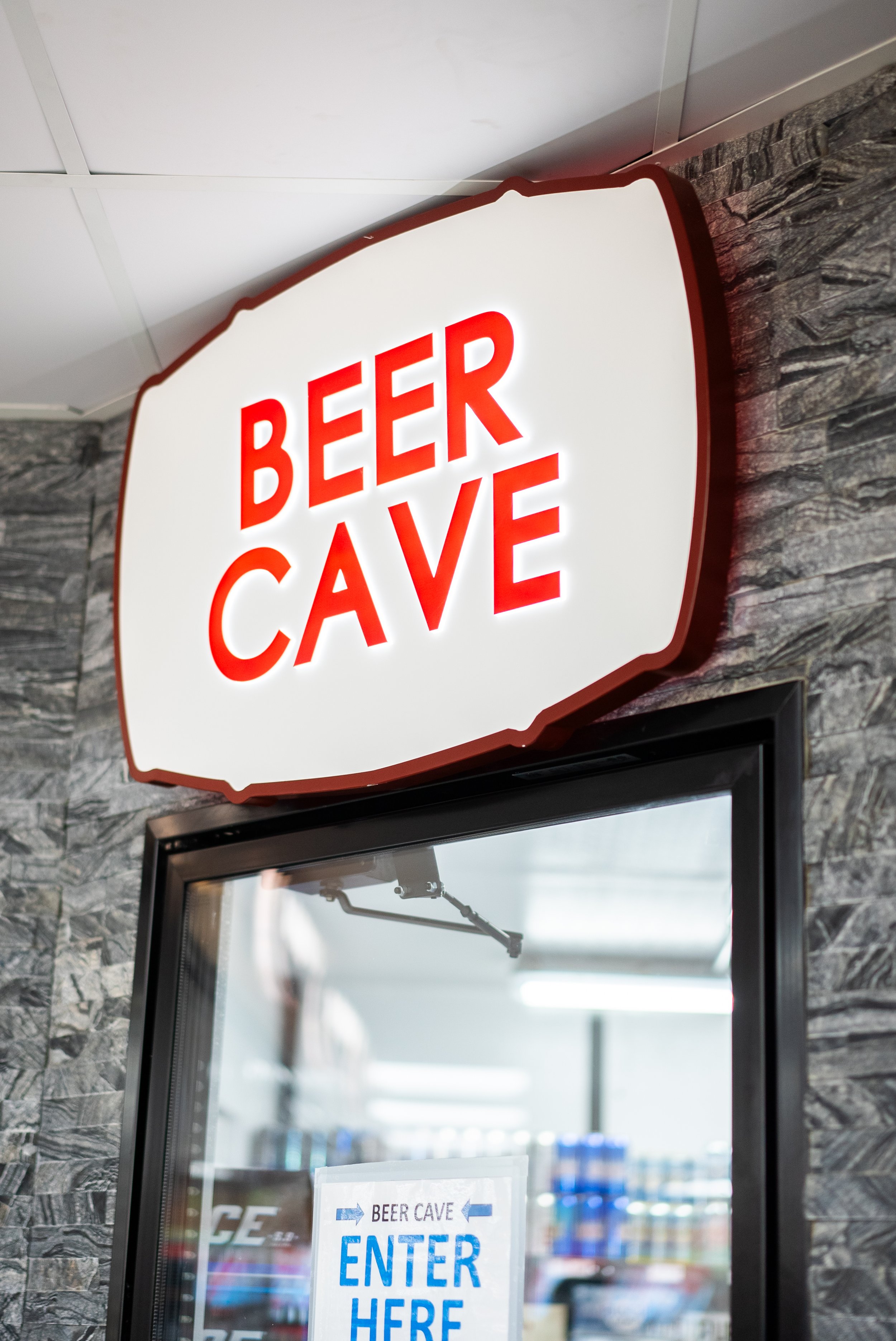 Bright illuminated sign reading "Beer Cave" mounted on a wall inside a store or bar, with a smaller sign below indicating "Enter Here".
