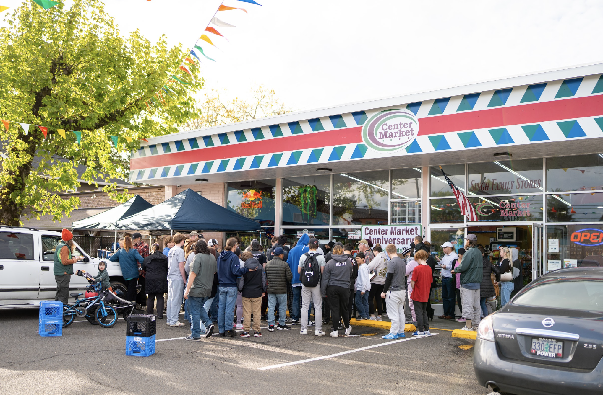 Crowd gathered outside Center Market for an event, with a line of people waiting to get inside on a parking lot, surrounded by trees and cars, under festive bunting and banners.