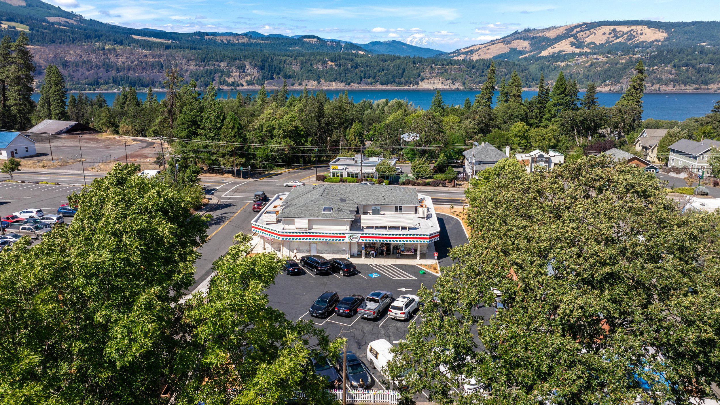 An aerial view of a small restaurant with a parking lot, surrounded by trees and a landscape of hills, a lake, and a sky with some clouds in the background.