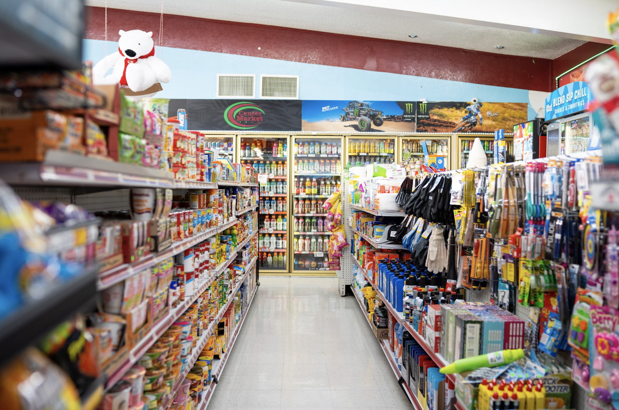 Aisle in a convenience store with shelves stocked with snacks, beverages, and miscellaneous items. A stuffed white dog with a red scarf is hanging from the ceiling, and coolers filled with drinks are visible in the background.