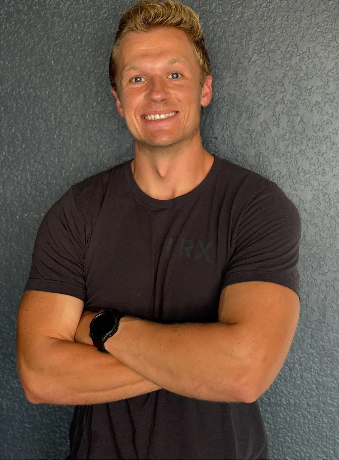 Smiling man with blond hair, wearing a black t-shirt, standing against a textured gray wall with arms crossed.