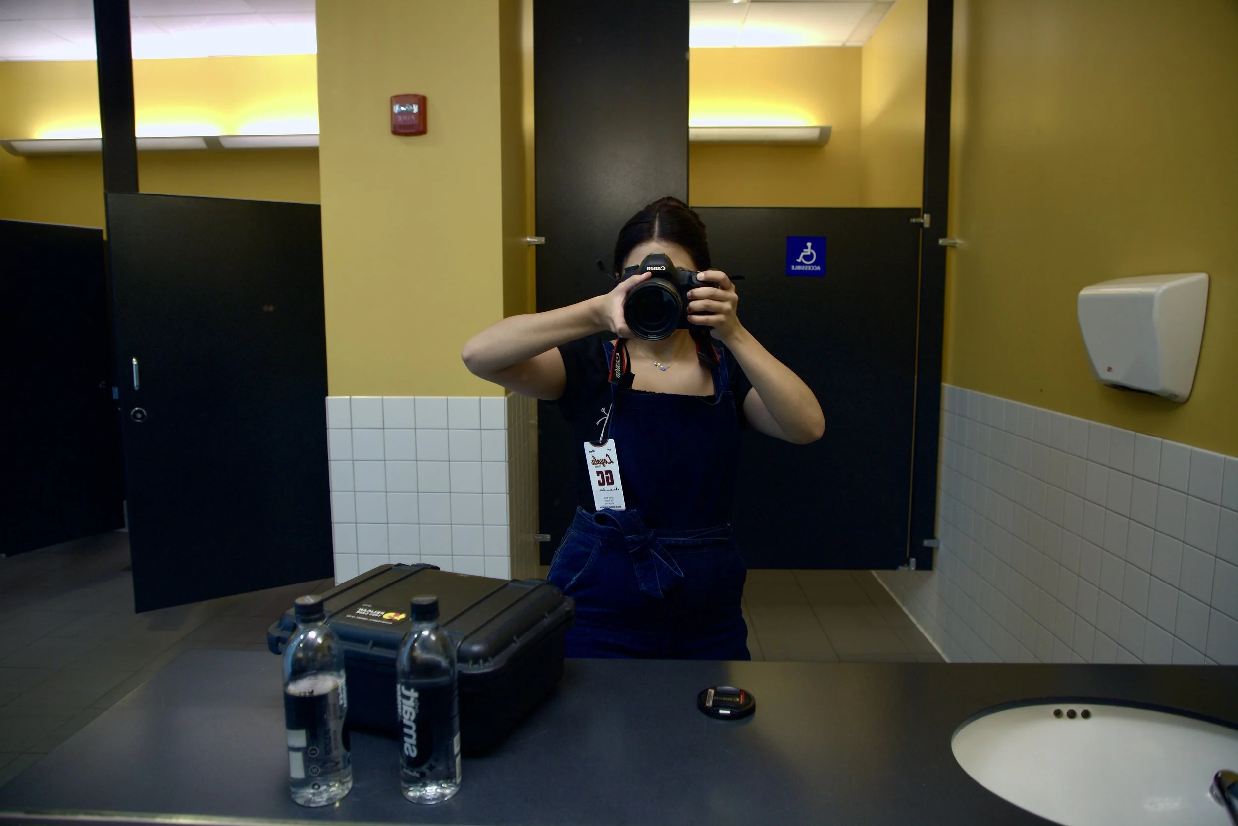 Person taking a mirror selfie in a restroom with a camera, wearing a lanyard and blue outfit. Two water bottles, a camera case, and a lens cap are on the counter.