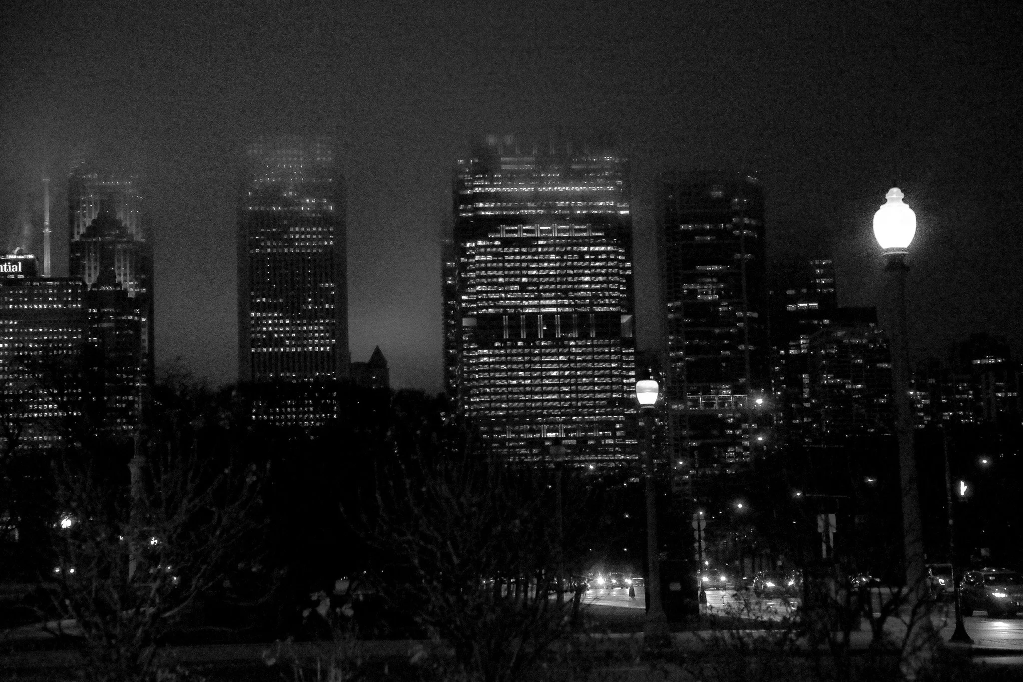 Black and white photo of a cityscape at night with tall buildings and bright street lamps.