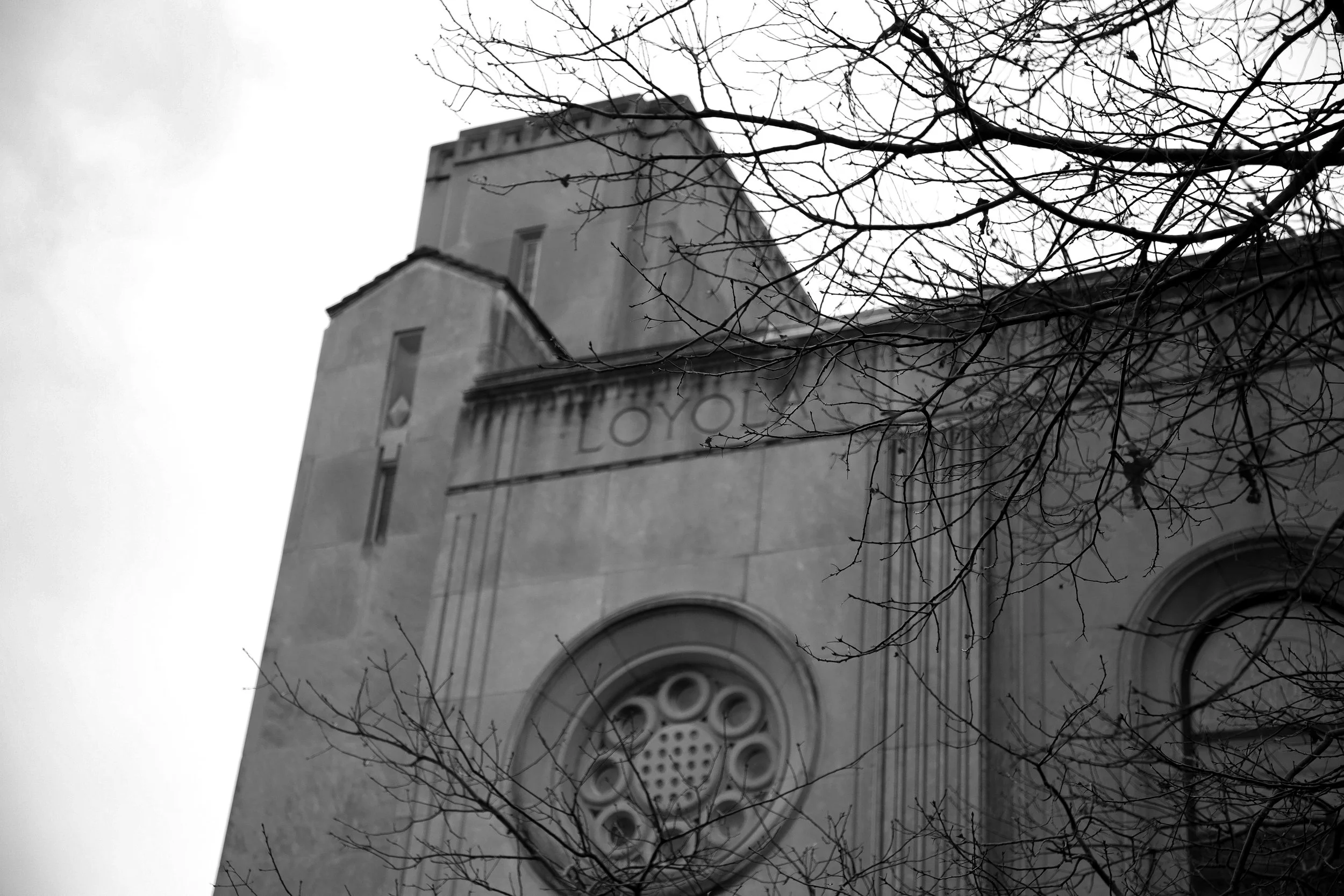 Black and white photo of a historic building with "Loyola" etched on the facade, featuring circular window designs and bare tree branches in the foreground.