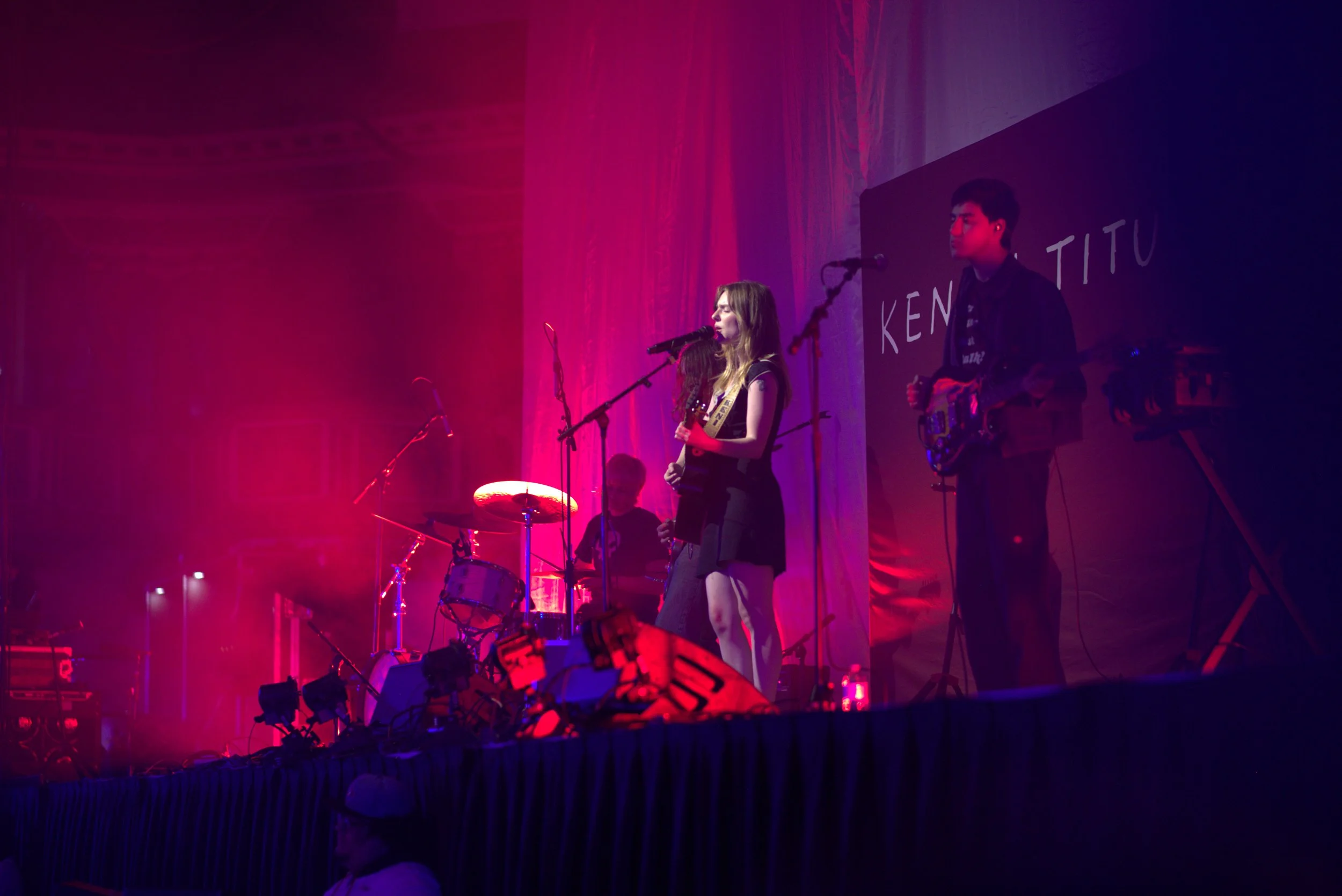 Band performing on stage with colorful lighting, featuring a female singer with a guitar, a drummer, and another musician playing a guitar. The stage is set with musical equipment, and there's a backdrop with partial text visible.