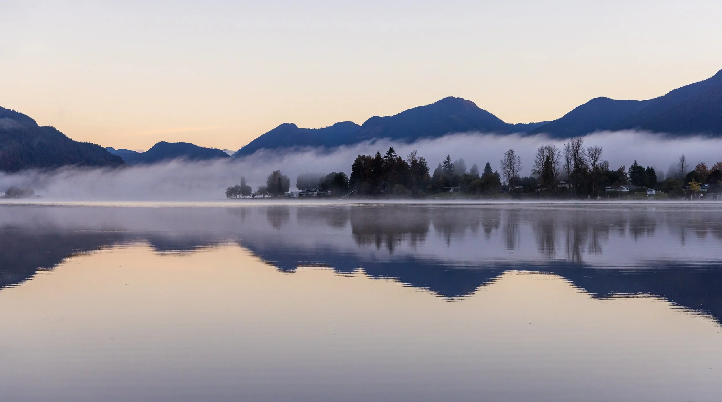 Lake with mist and trees reflected in water; mountains in background
