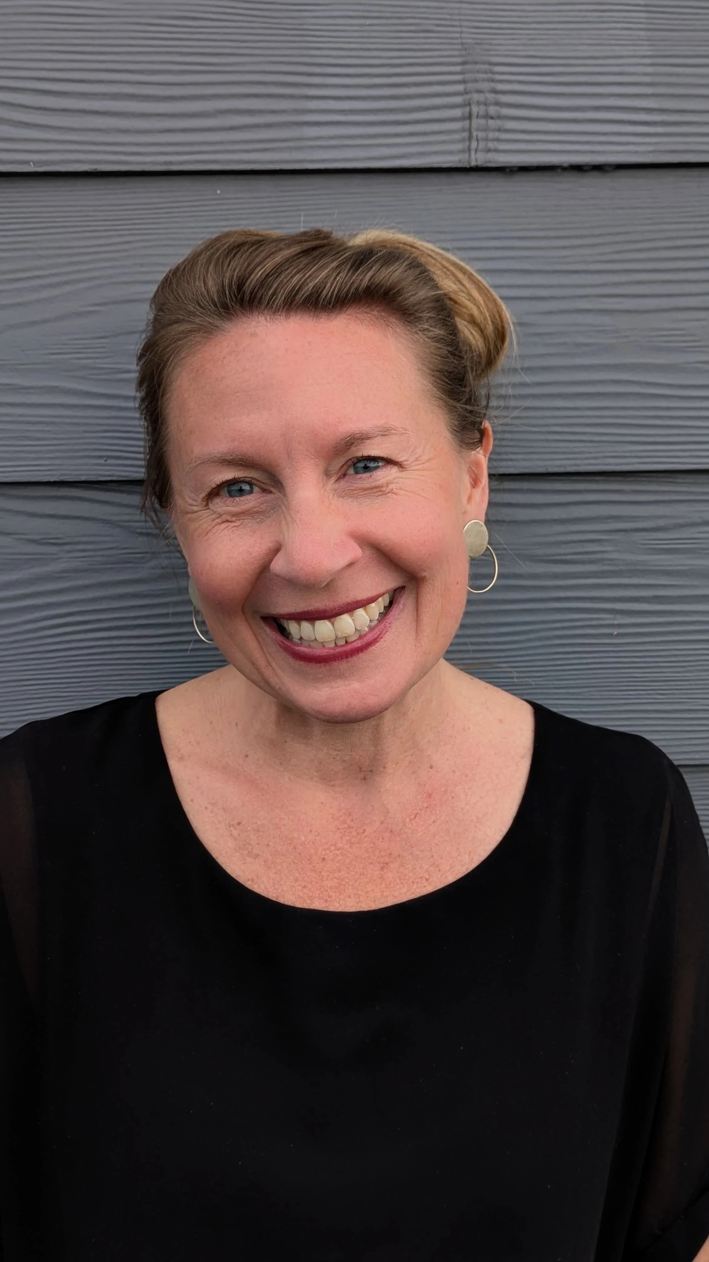 A woman with short hair is smiling, wearing a black top and circular earrings, standing against a gray wooden wall.