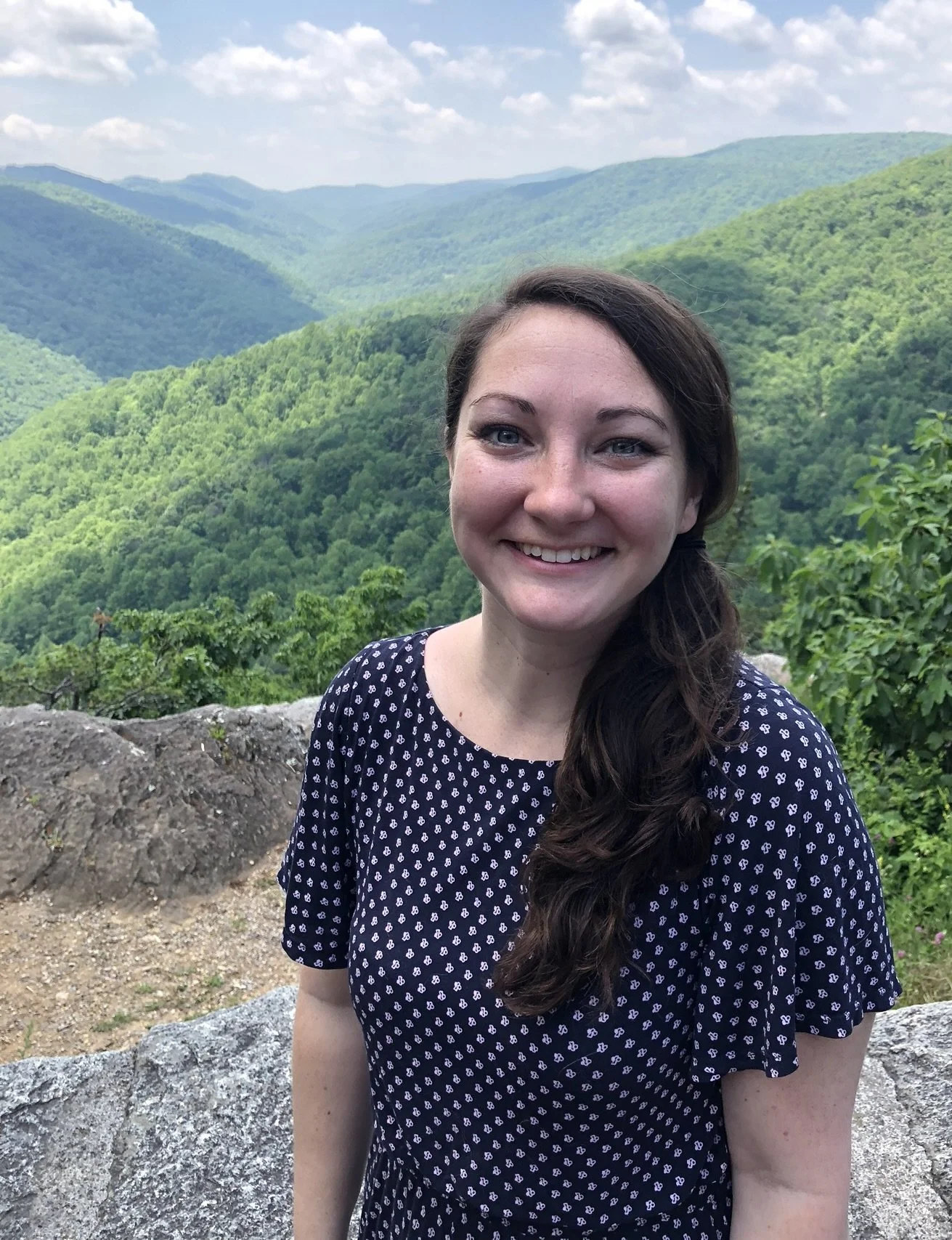Smiling woman in a navy blue dress with white pattern standing outdoors with green mountain landscape in the background.
