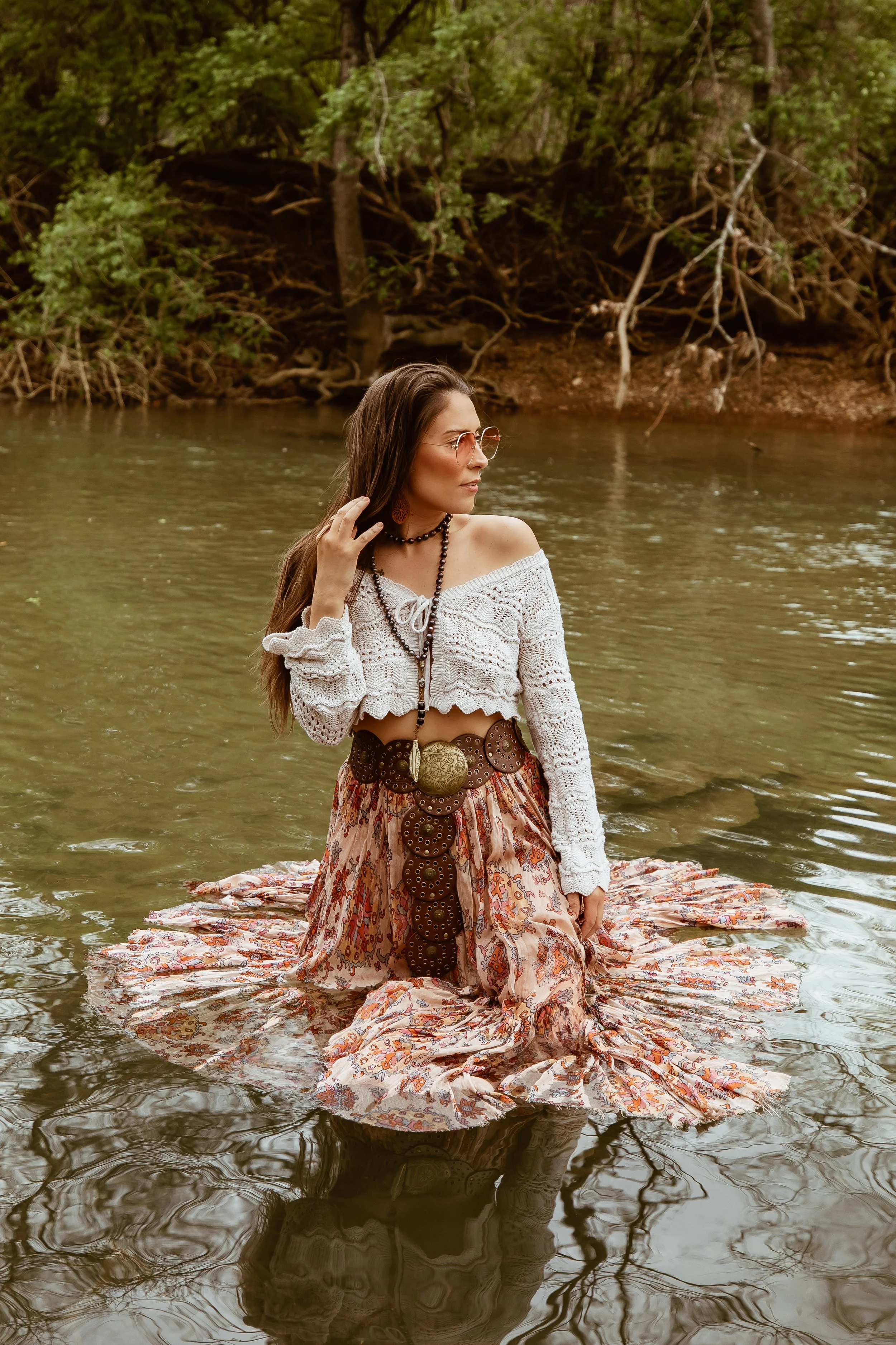 Woman in bohemian-style outfit standing in a shallow river, wearing a white crochet top and floral skirt. She is touching her hair and surrounded by green trees.
