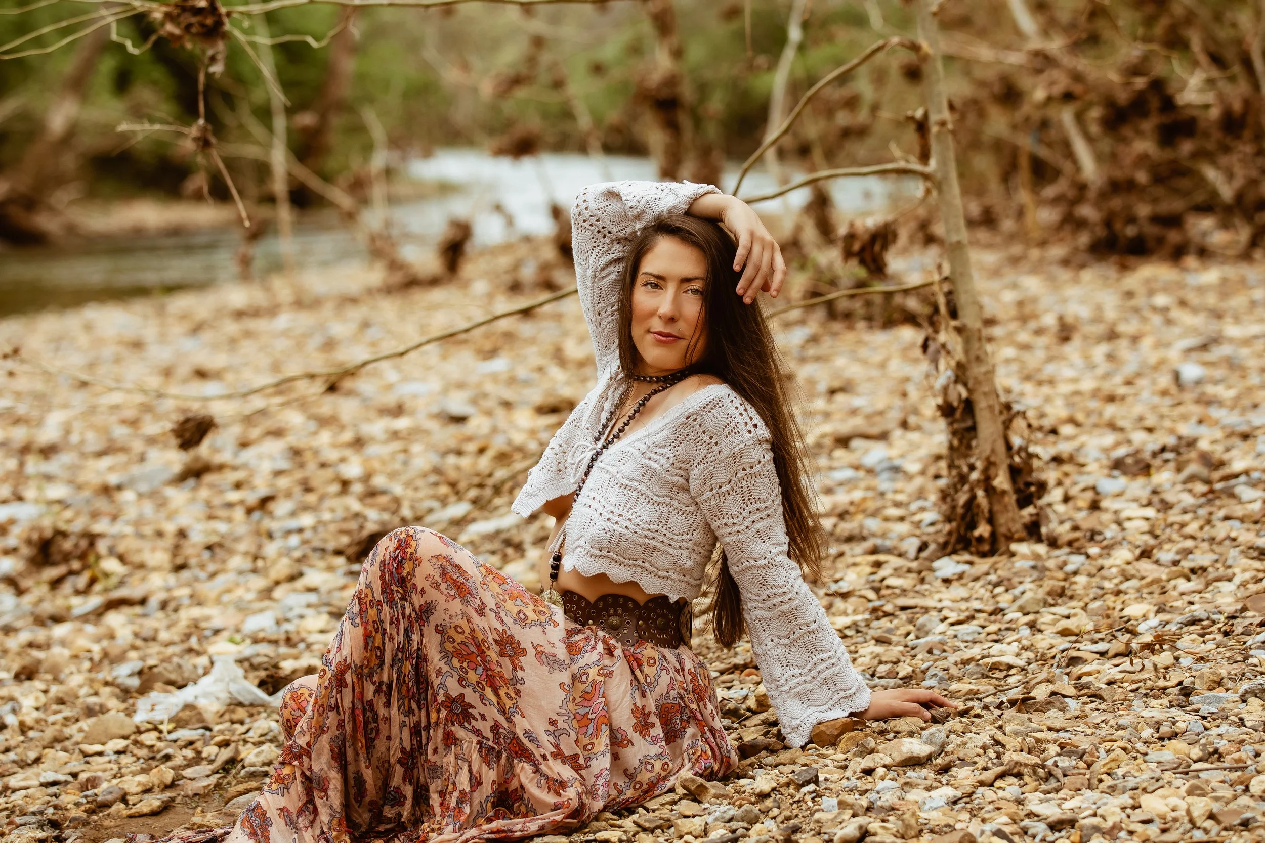 Woman sitting on rocky ground near a stream, wearing a white crocheted top and a floral skirt, surrounded by trees and natural scenery.