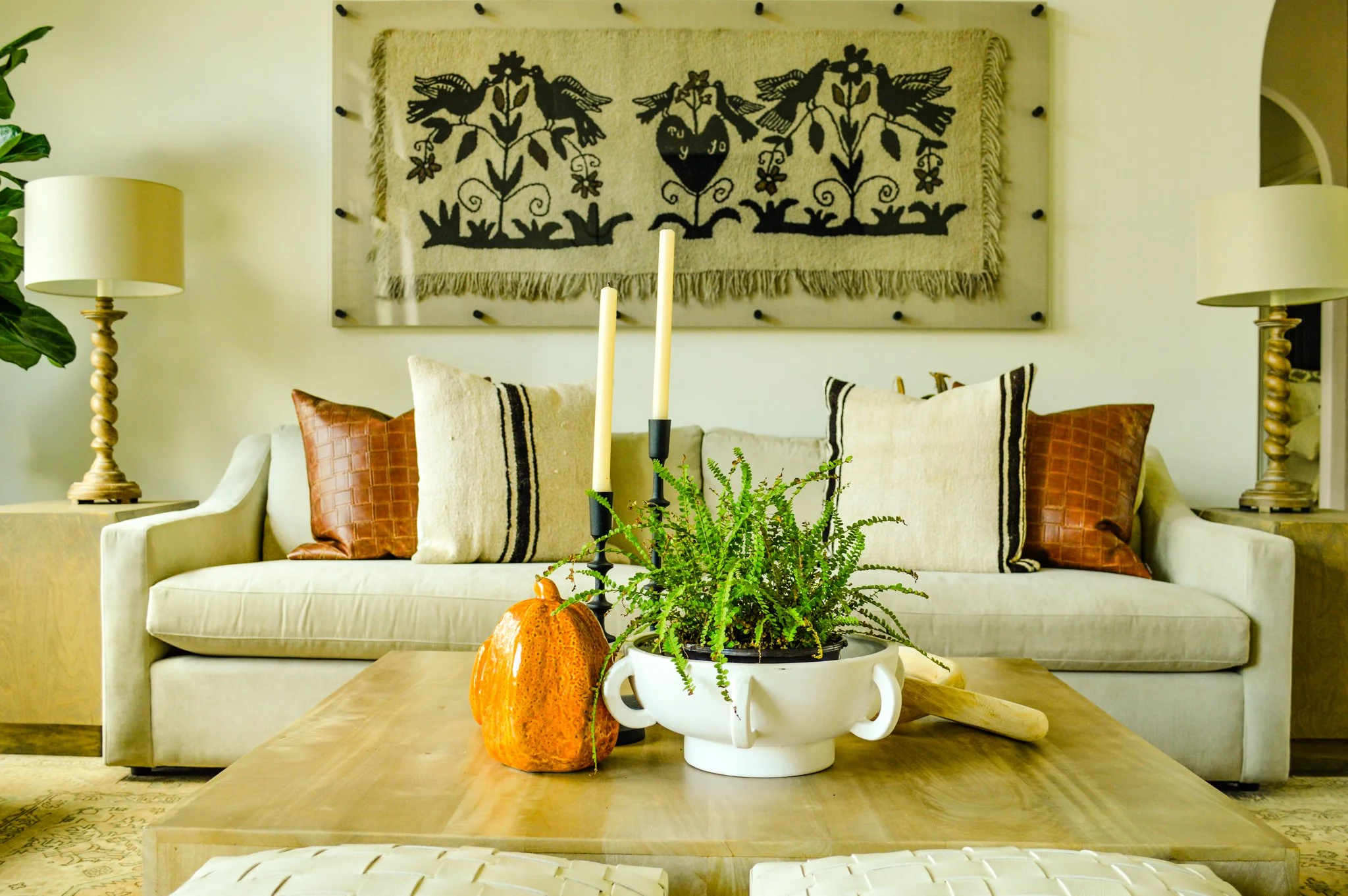 A living room with a beige sofa decorated with brown and beige pillows. A large wall hanging with black floral and bird patterns is mounted above the sofa. There are two white table lamps with twisted bases on side tables on either side of the sofa, a potted fern in a white pot and an orange decorative pumpkin on a wooden coffee table in front of the sofa. A large green plant is visible on the left side.