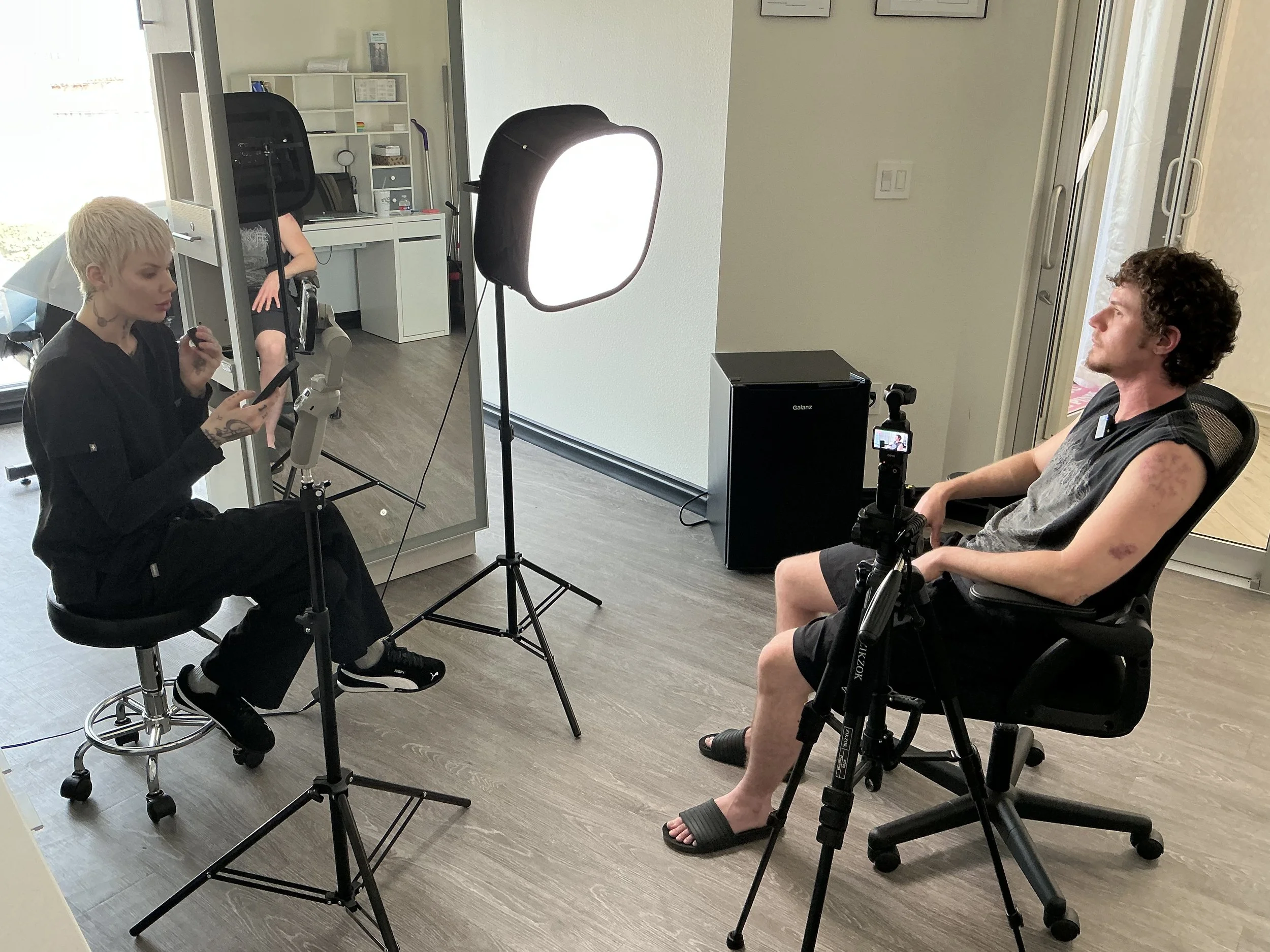 A woman and a man are sitting in a room with photography or filming equipment around them. The woman is reading from a phone, and the man is sitting in a chair facing her. There is a large softbox light directed towards the sitting man, a camera on a tripod, and a reflective surface. The background includes a white desk and shelving with small items.