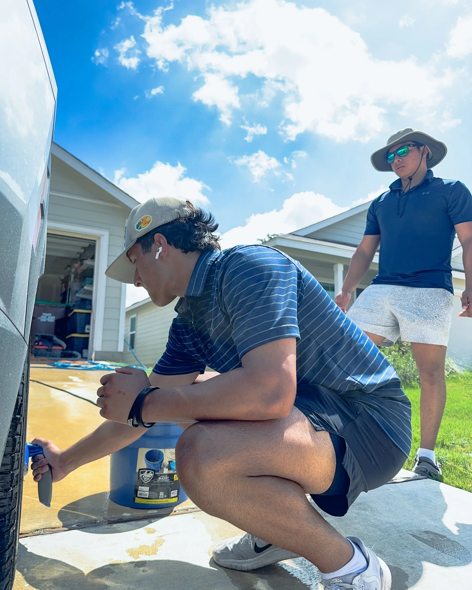 Two men working outside on a sunny day, one kneeling and using a tool near a utility box, and the other standing nearby wearing sunglasses and a hat, with a house in the background.