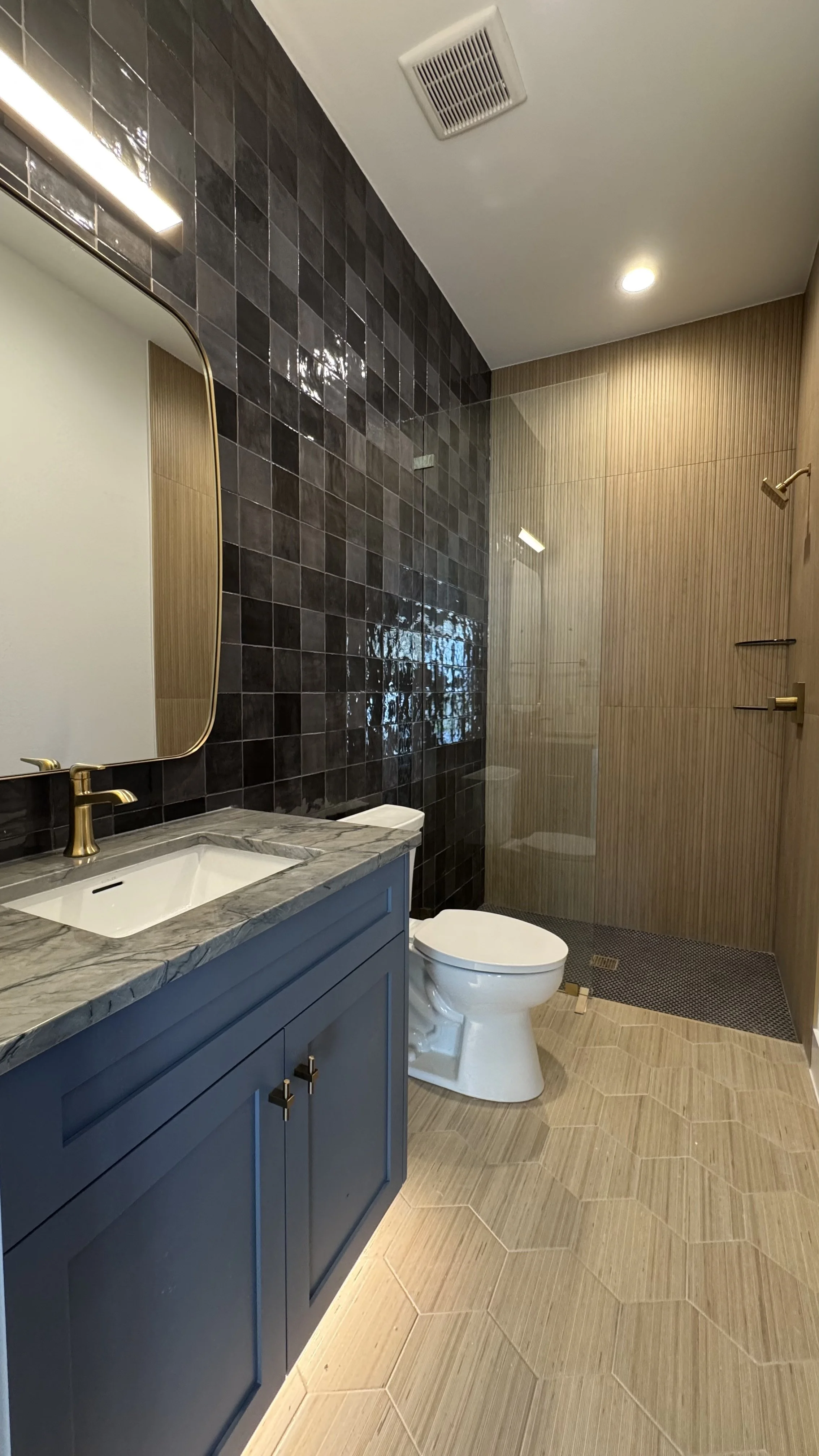 Modern bathroom with a blue vanity, marble countertop, mirror, dark tiled wall, beige tiled shower area with glass door, white toilet, ceiling vent, and recessed lighting.