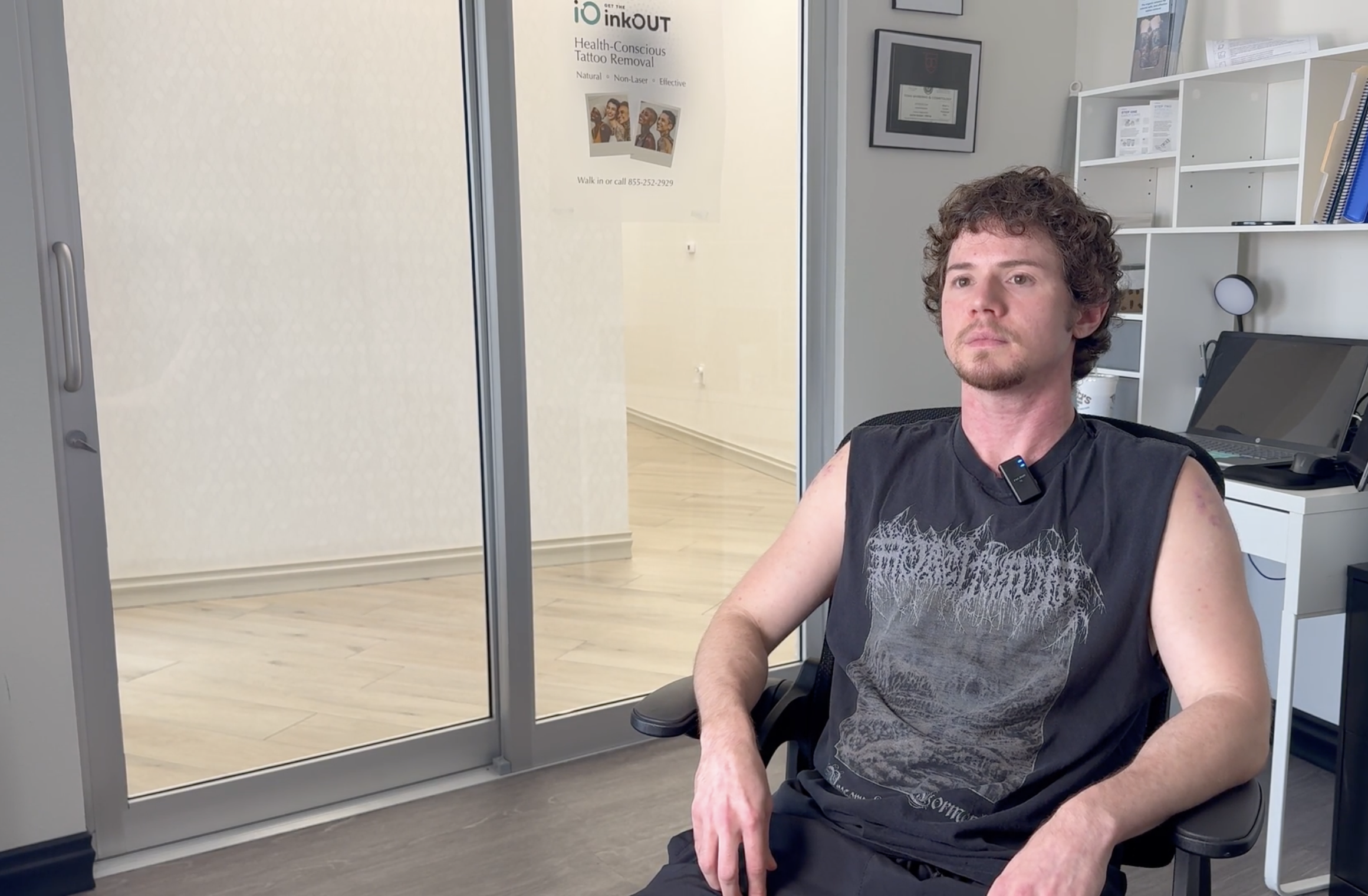 A man with curly hair and a beard sitting in a black chair, wearing a sleeveless black T-shirt, in a medical or office setting, with a white shelving unit and framed certificate or diploma on the wall behind him, and a poster about tattoo removal on the glass door.