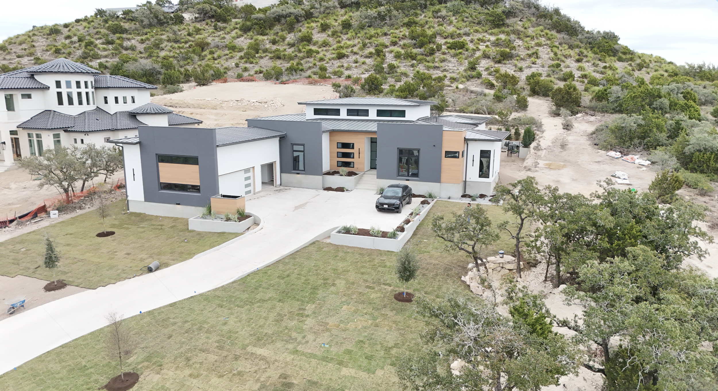 An aerial view of a modern house under construction with a white and gray exterior, large windows, a driveway with a parked car, a grassy yard, and a hillside with trees in the background.
