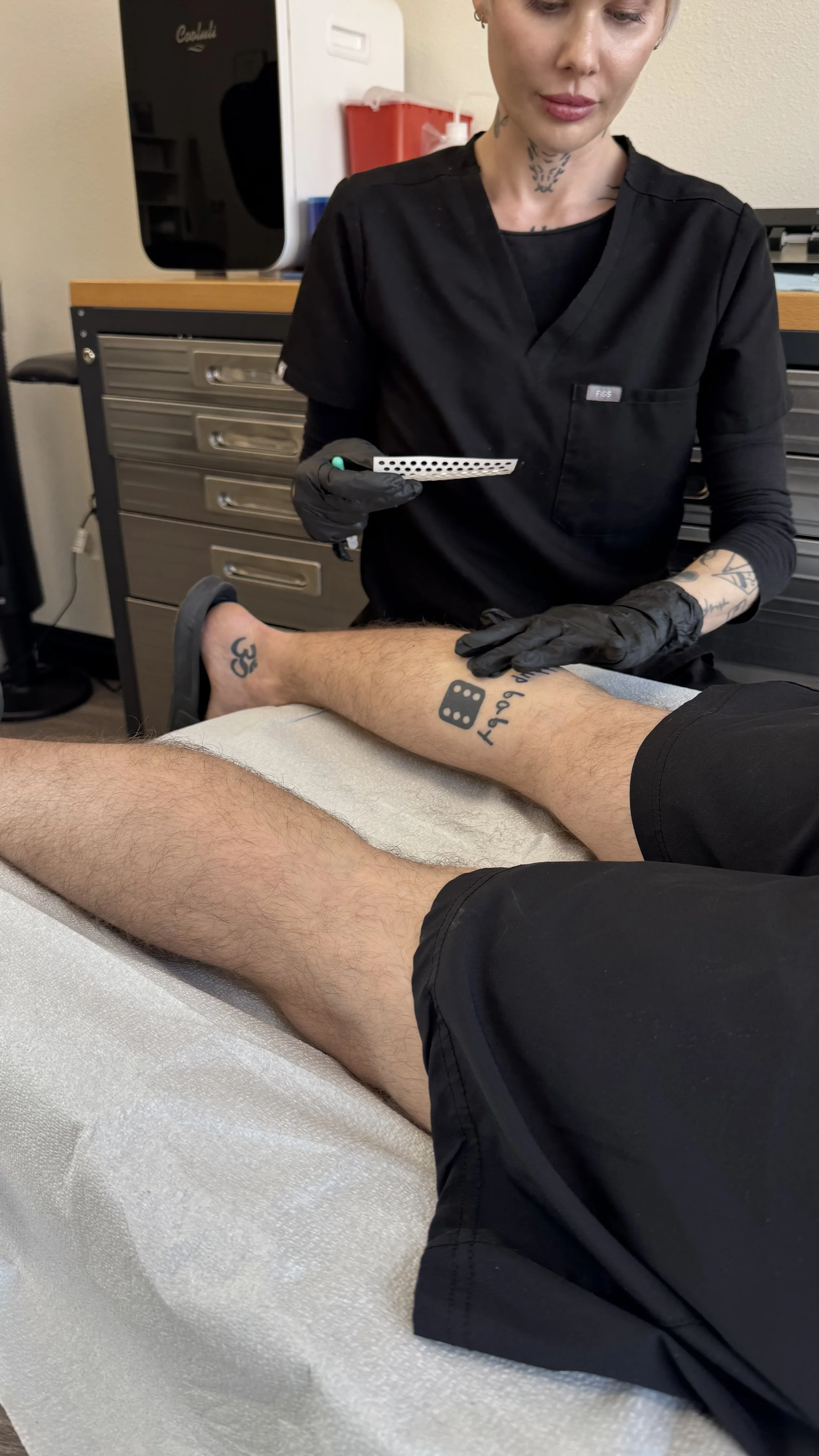 A healthcare professional wearing black scrubs and gloves drawing blood from a patient's leg vein using a needle and tube in a medical setting.