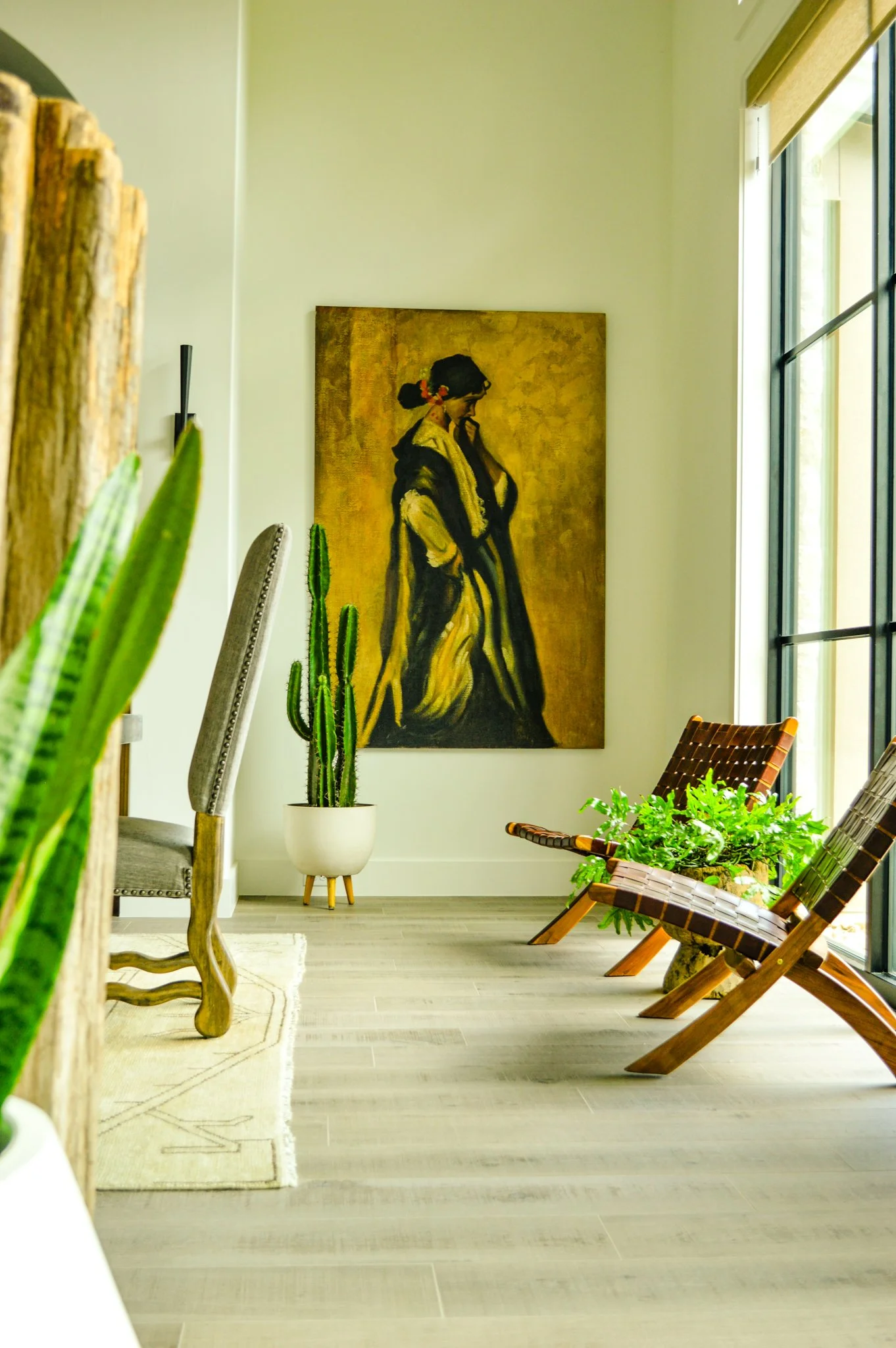 Interior of a room with a large window on the right, a painting of a woman on the wall, potted plants including a cactus, and wooden chairs with woven seats.