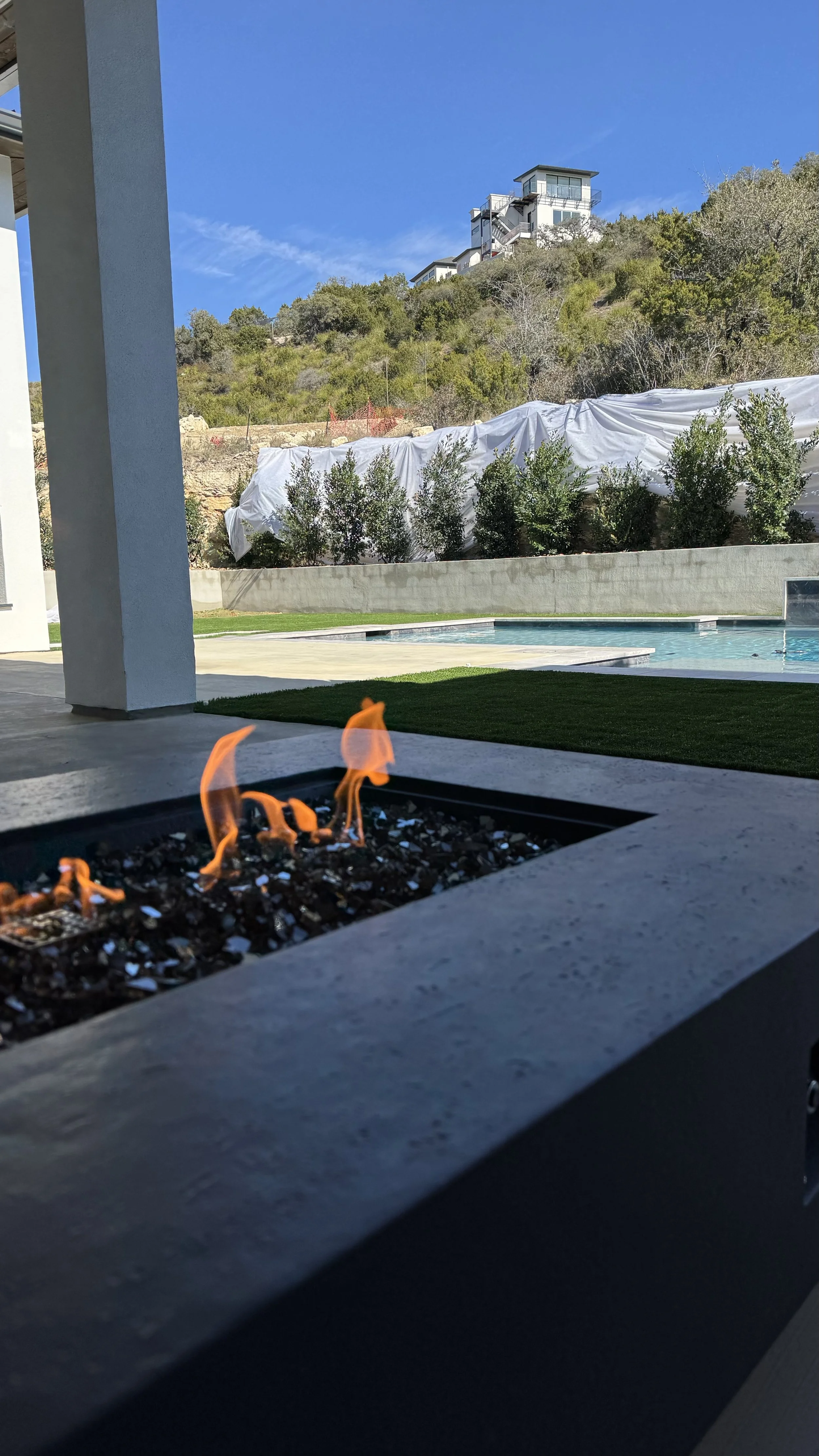 A modern backyard with a fire pit, swimming pool, green lawn, and hillside with trees and houses under a clear blue sky.