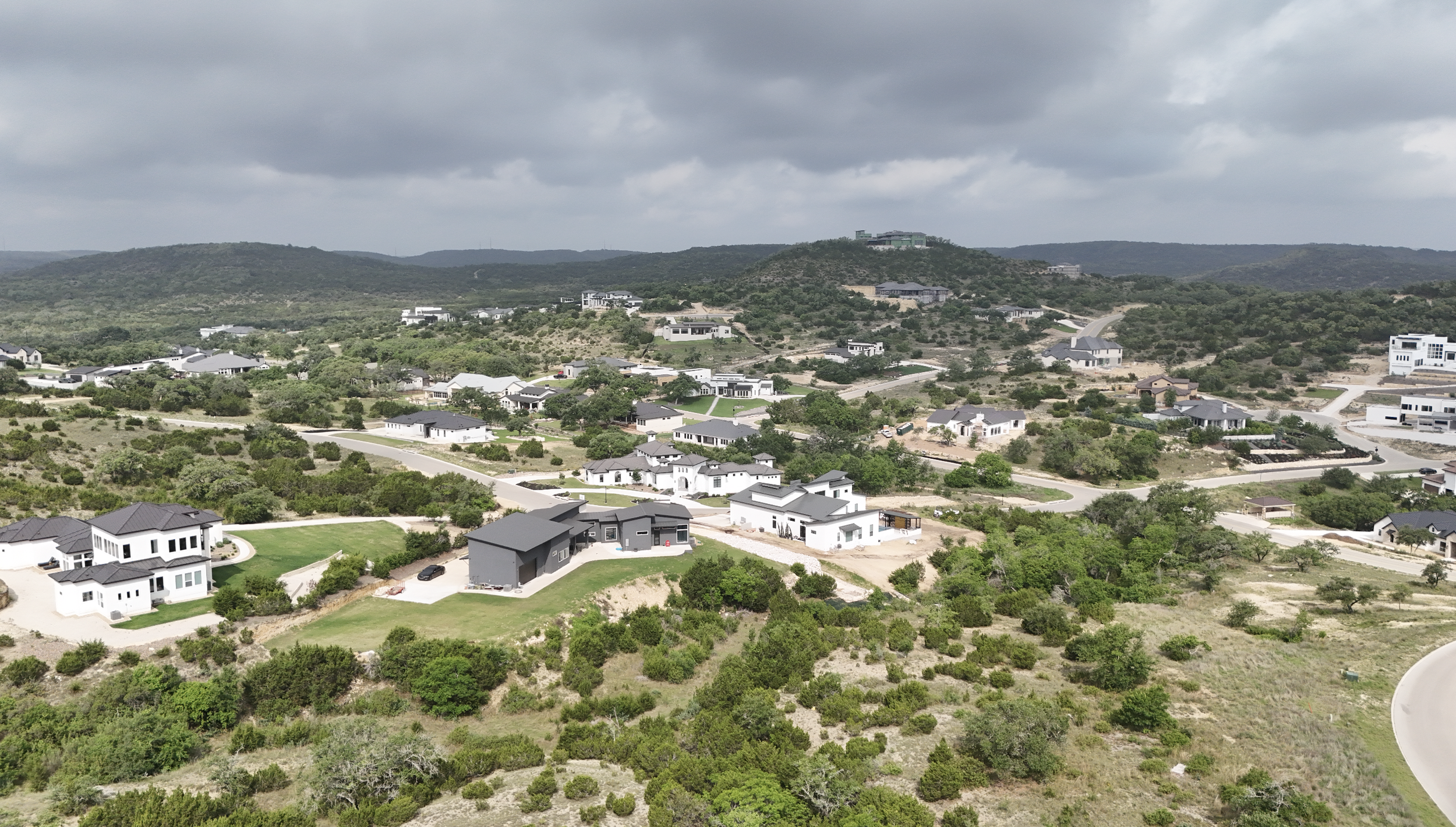 An aerial view of a suburban neighborhood with houses, roads, and green vegetation, set against hilly terrain and a cloudy sky.