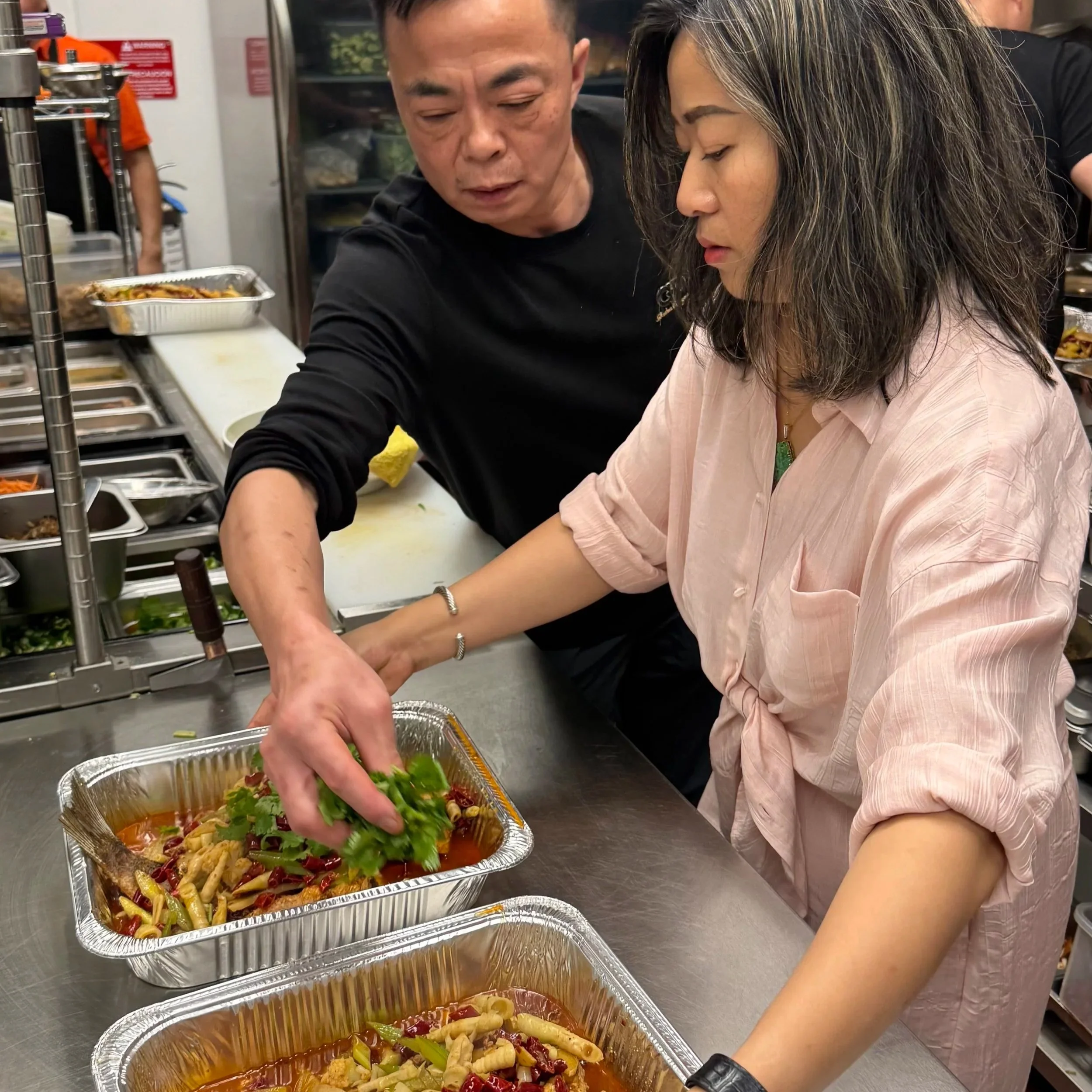 Two people preparing food in a restaurant kitchen, with one woman placing salad into a foil container and a man assisting.