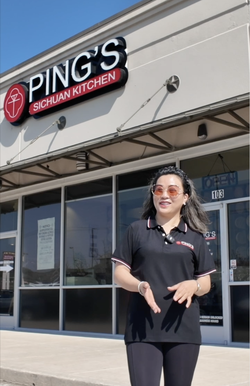 A woman standing outside a restaurant called Ping's Sichuan Kitchen, smiling and gesturing with her hands, wearing sunglasses and a black uniform.