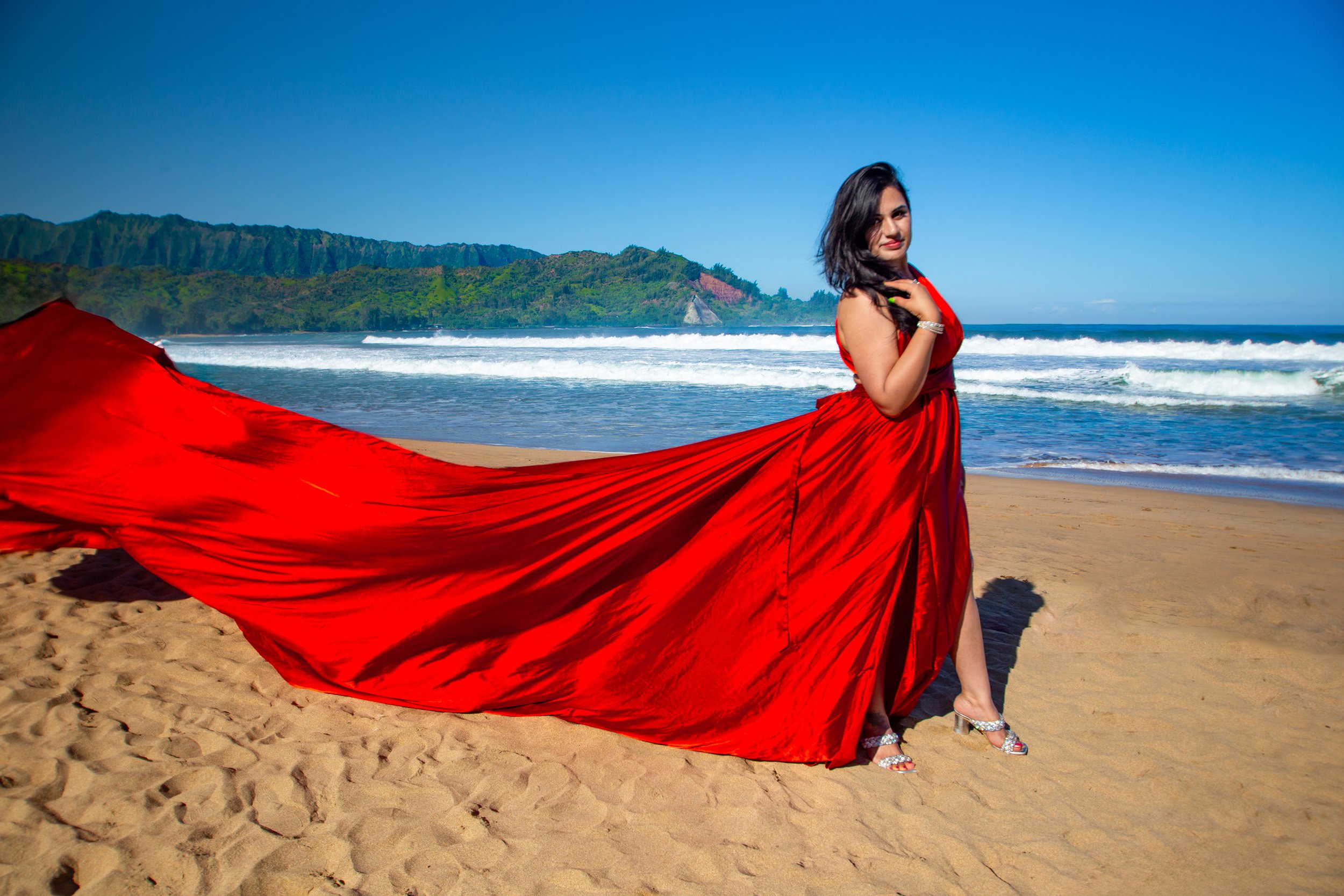 A woman in a red gown standing on a sandy beach with ocean waves and green hills in the background.