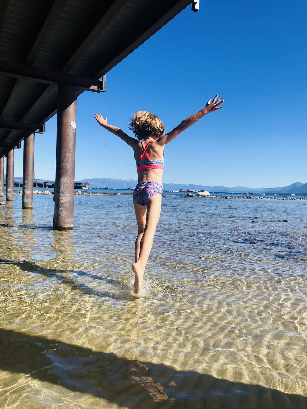 A young girl in a swimsuit jumping in shallow water under a pier at the beach with mountains in the background.