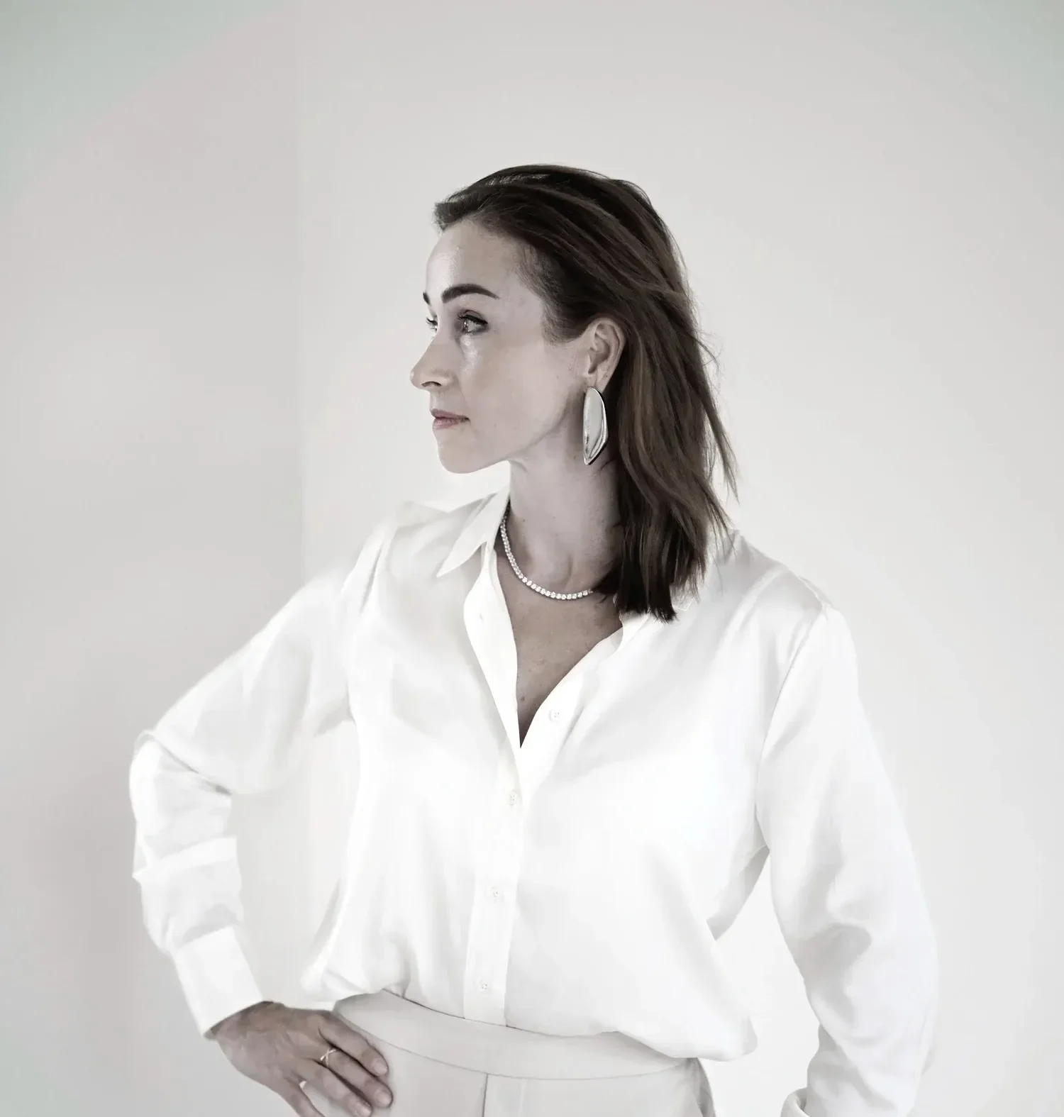 A woman with dark hair in a profile view wearing a white button-down shirt, a tennis necklace, and silver statement earrings.