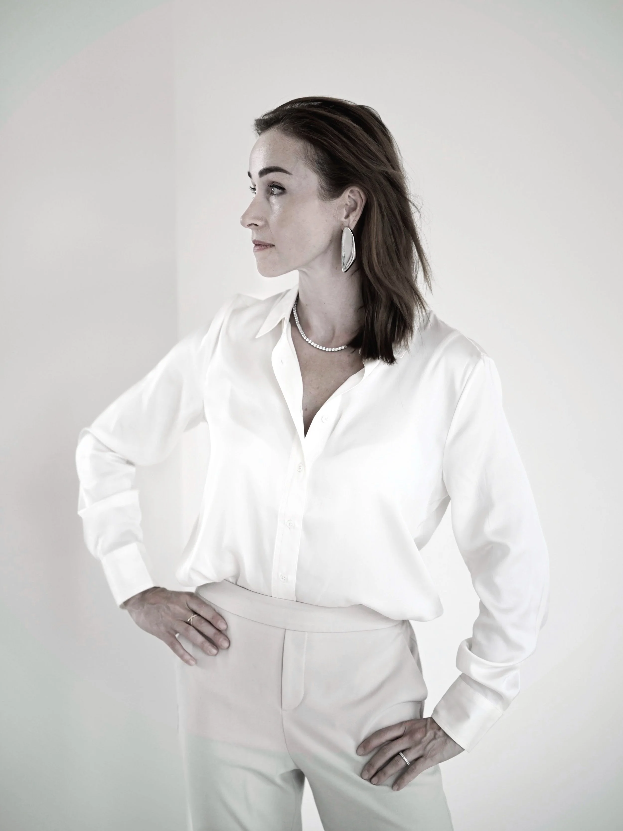 A woman with dark brown hair wearing a white blouse and cream-colored trousers, standing with hands on hips and looking to the side against a plain, light-colored background.