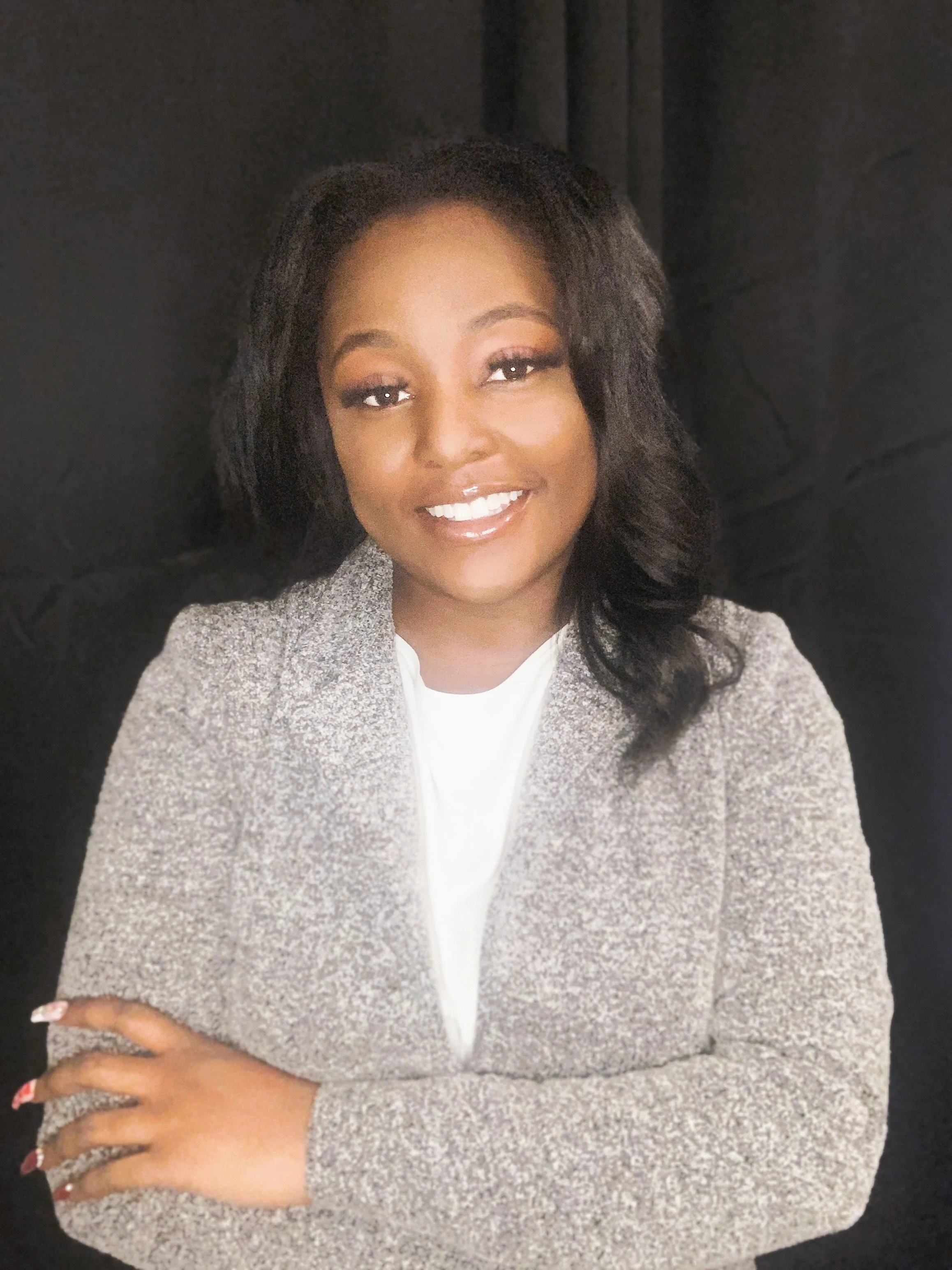 A woman with dark hair, smiling, wearing a gray blazer and white top, against a black background.