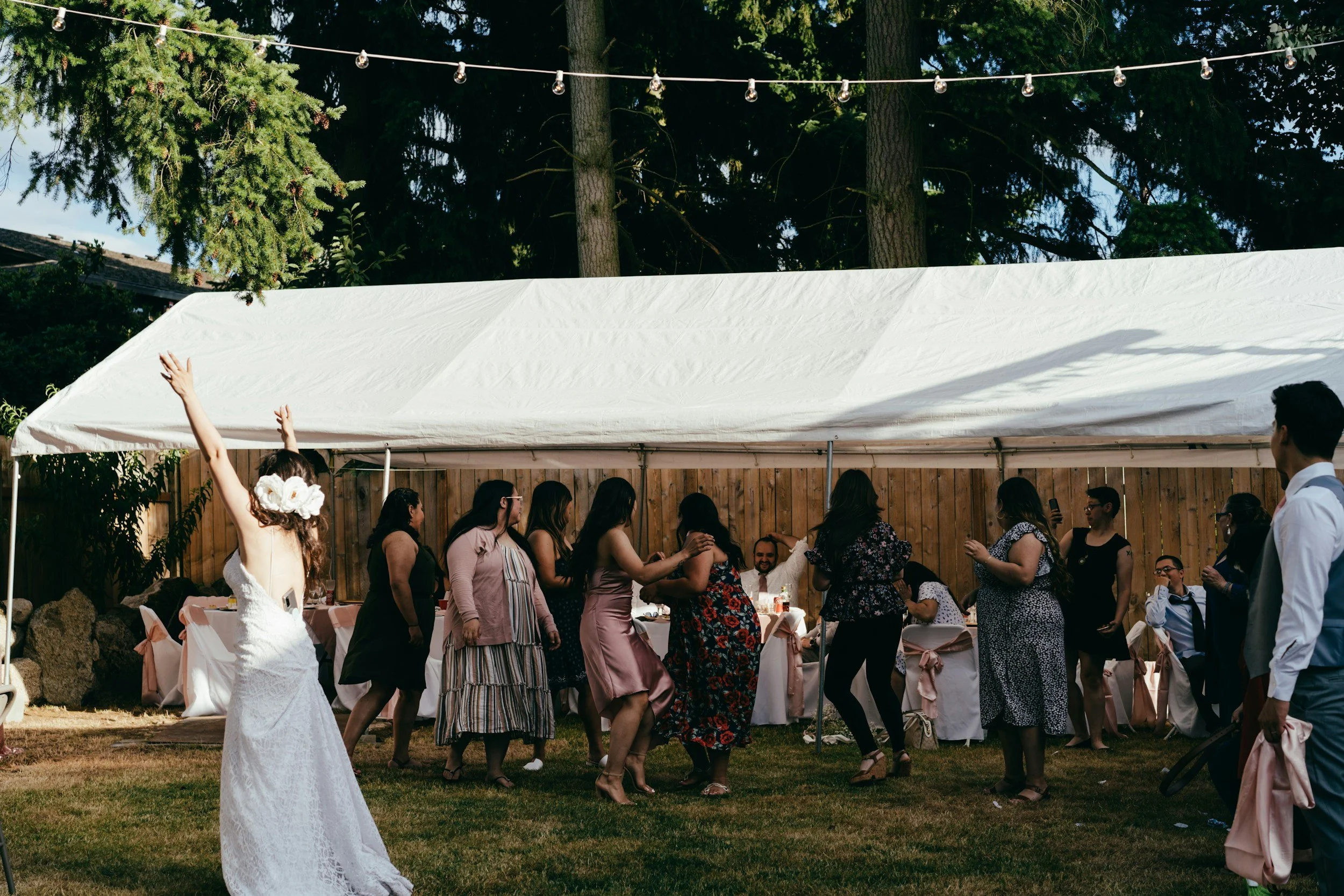A group of people dancing and socializing outdoors during a celebration, with a white canopy tent and string lights in the background.