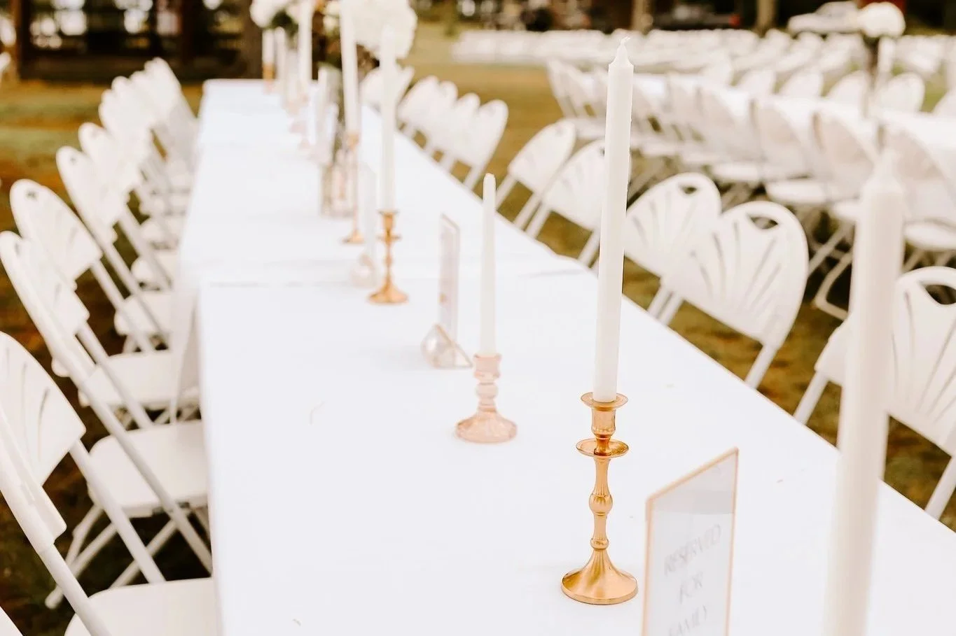 Set up for an outdoor event with white chairs and long tables decorated with white and gold candles, and a reserved sign on the table.