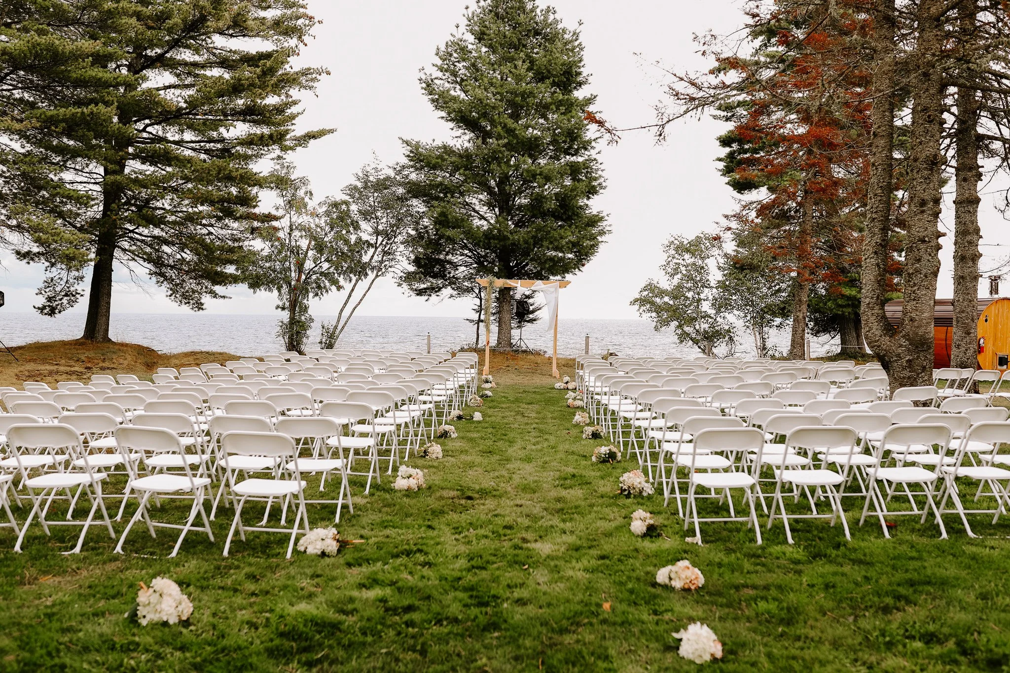 Outdoor wedding ceremony setup with white chairs aligned on grass facing an arch, decorated with flowers, under tall trees near a body of water.