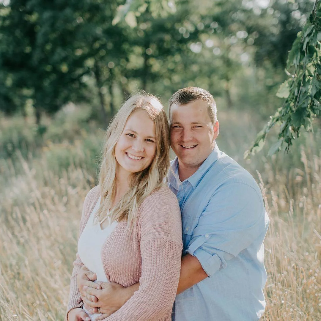 A smiling couple standing close together outdoors among tall grasses and trees, with the man embracing the woman from behind.