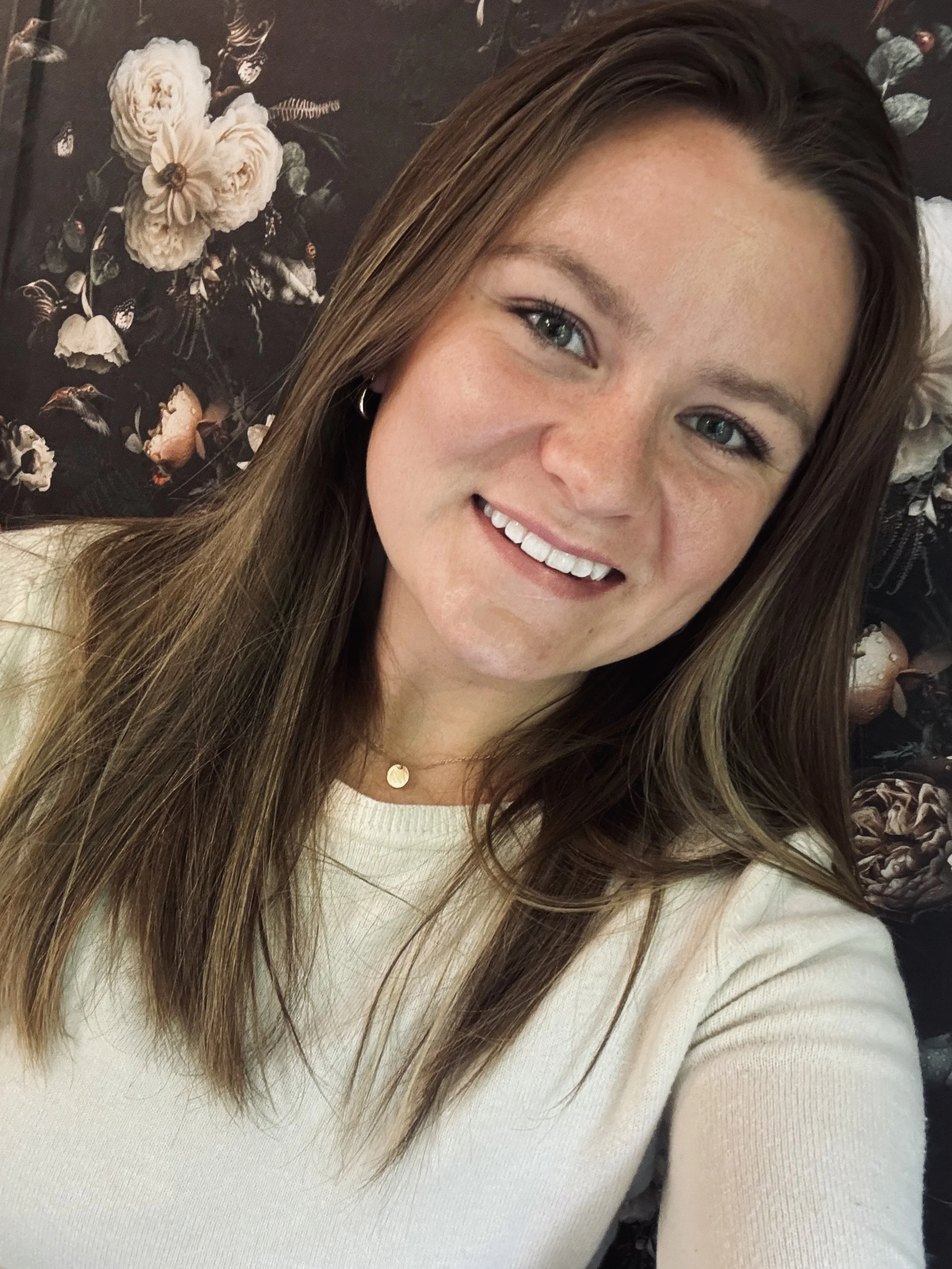 A young woman with long brown hair, blue eyes, and a bright smile, wearing a white top and a delicate gold necklace with a small pendant, taking a selfie in front of a dark floral background with beige flowers.