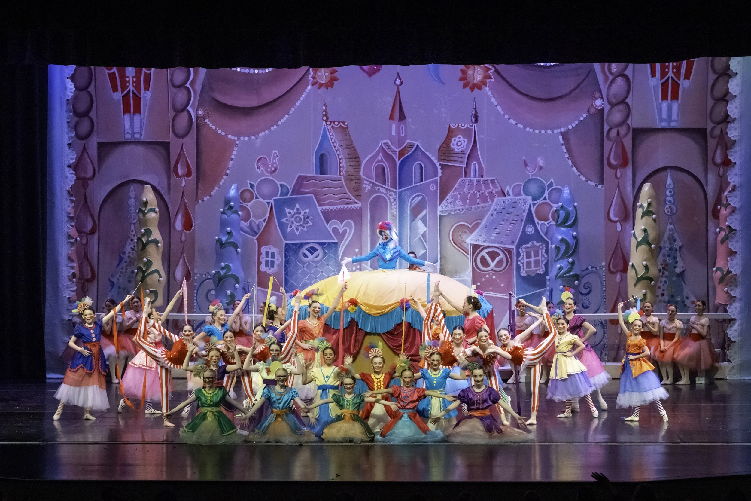 Children in colorful costumes performing on stage with a whimsical backdrop, featuring a princess at the center and dancers with striped and solid-colored dresses.