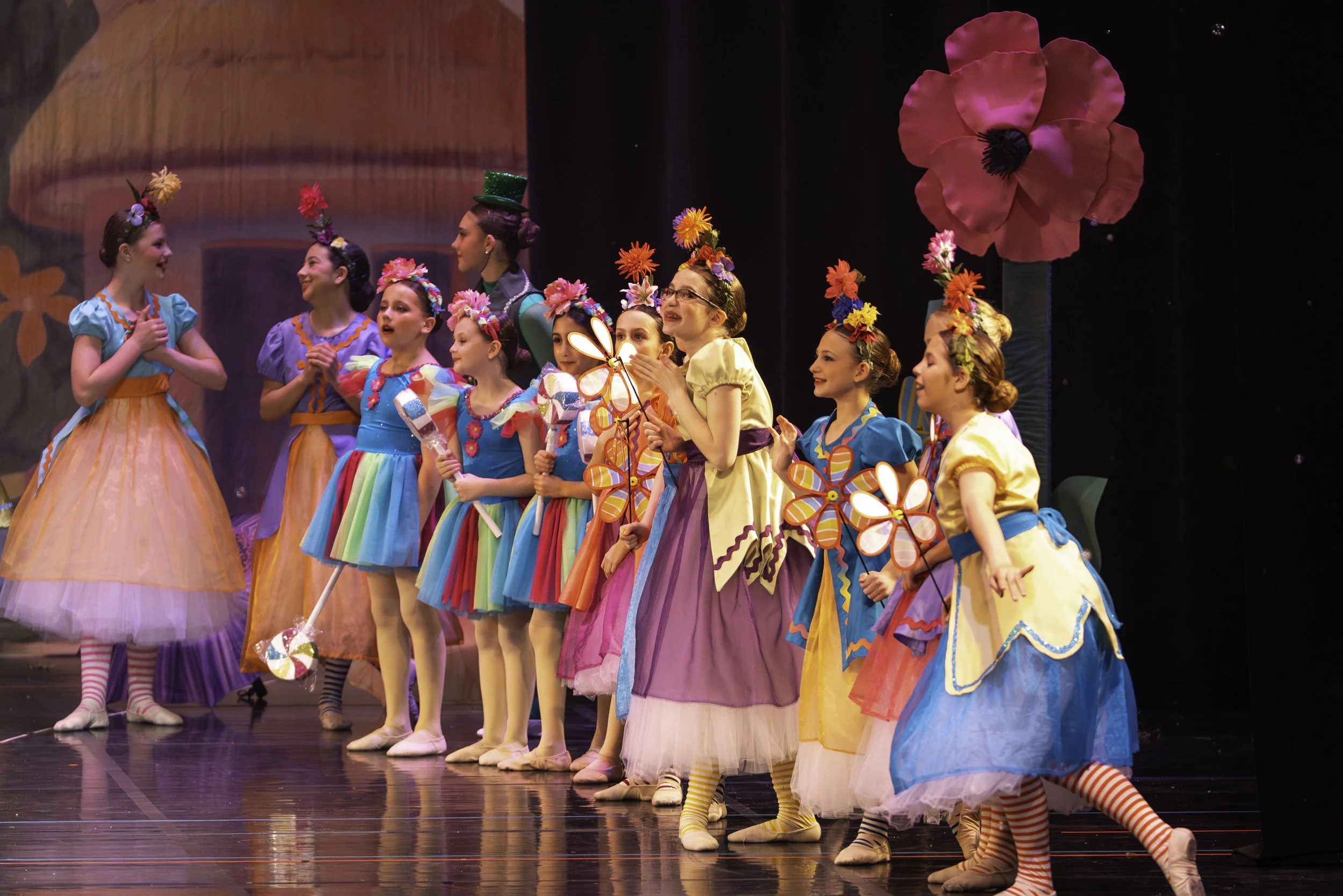 A group of young girls dressed in colorful costumes and flower accessories on stage, taking a bow after a dance performance.