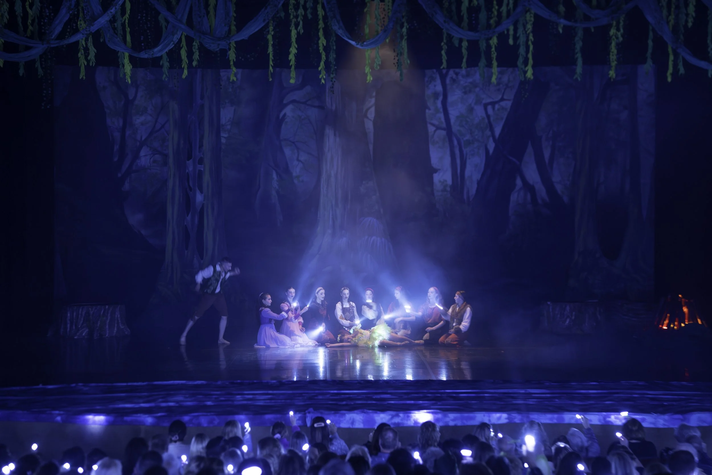 People dressed in costumes performing a theatrical scene on a stage with a forest backdrop, illuminated with blue stage lights and surrounded by an audience.