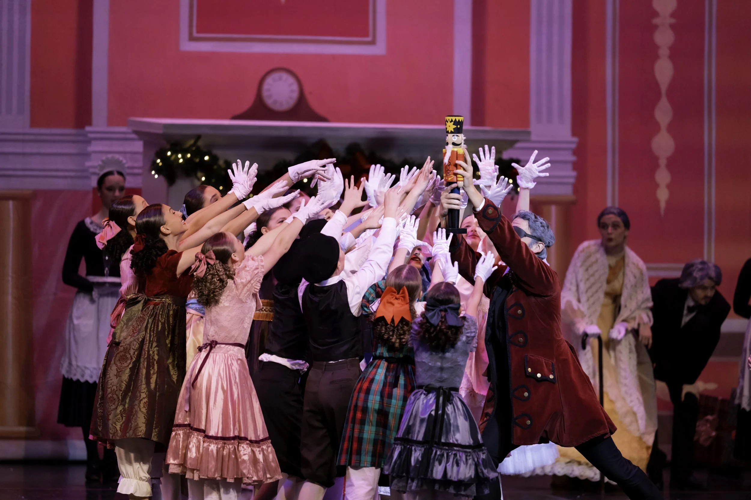 Children and adults in Victorian costumes reaching towards a toy soldier on a stage with pink walls and Christmas decorations.